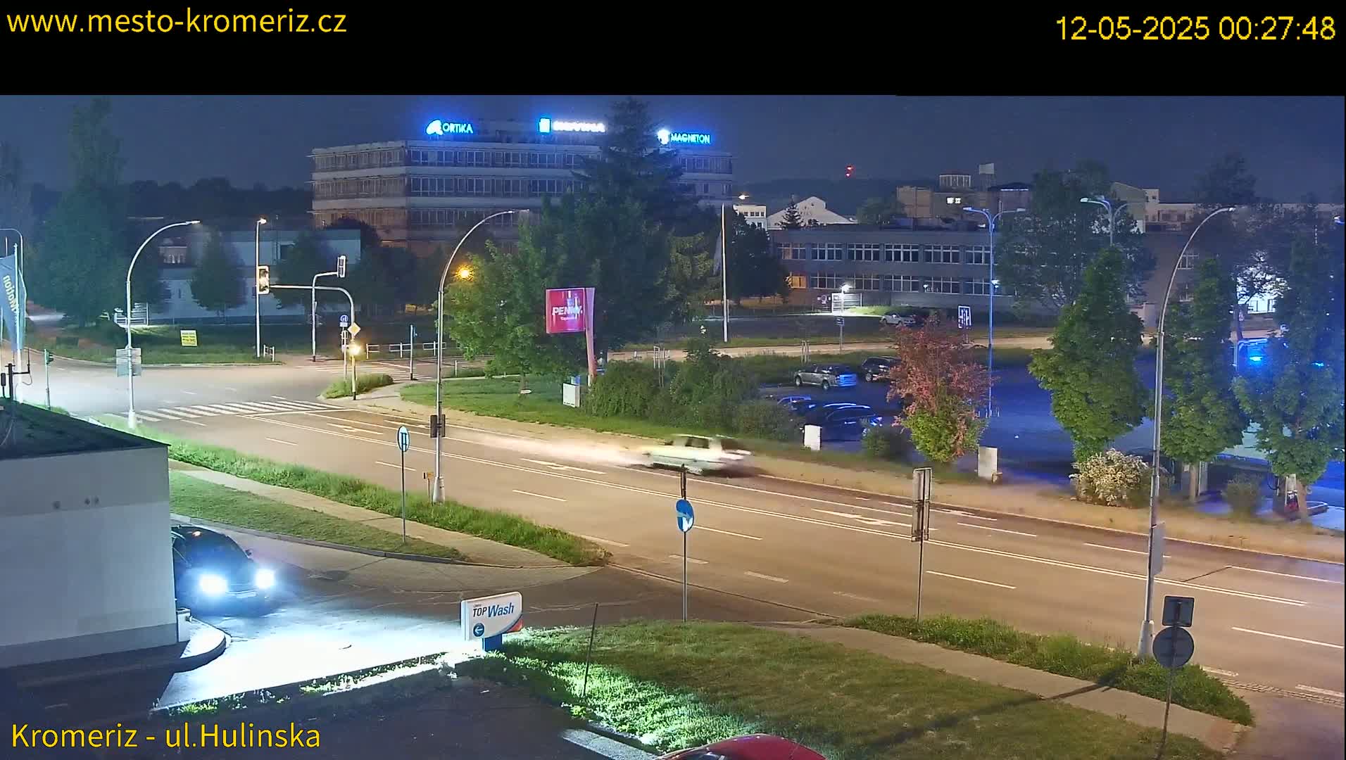 A nighttime view of a multi-lane road with cars driving past buildings under streetlights.