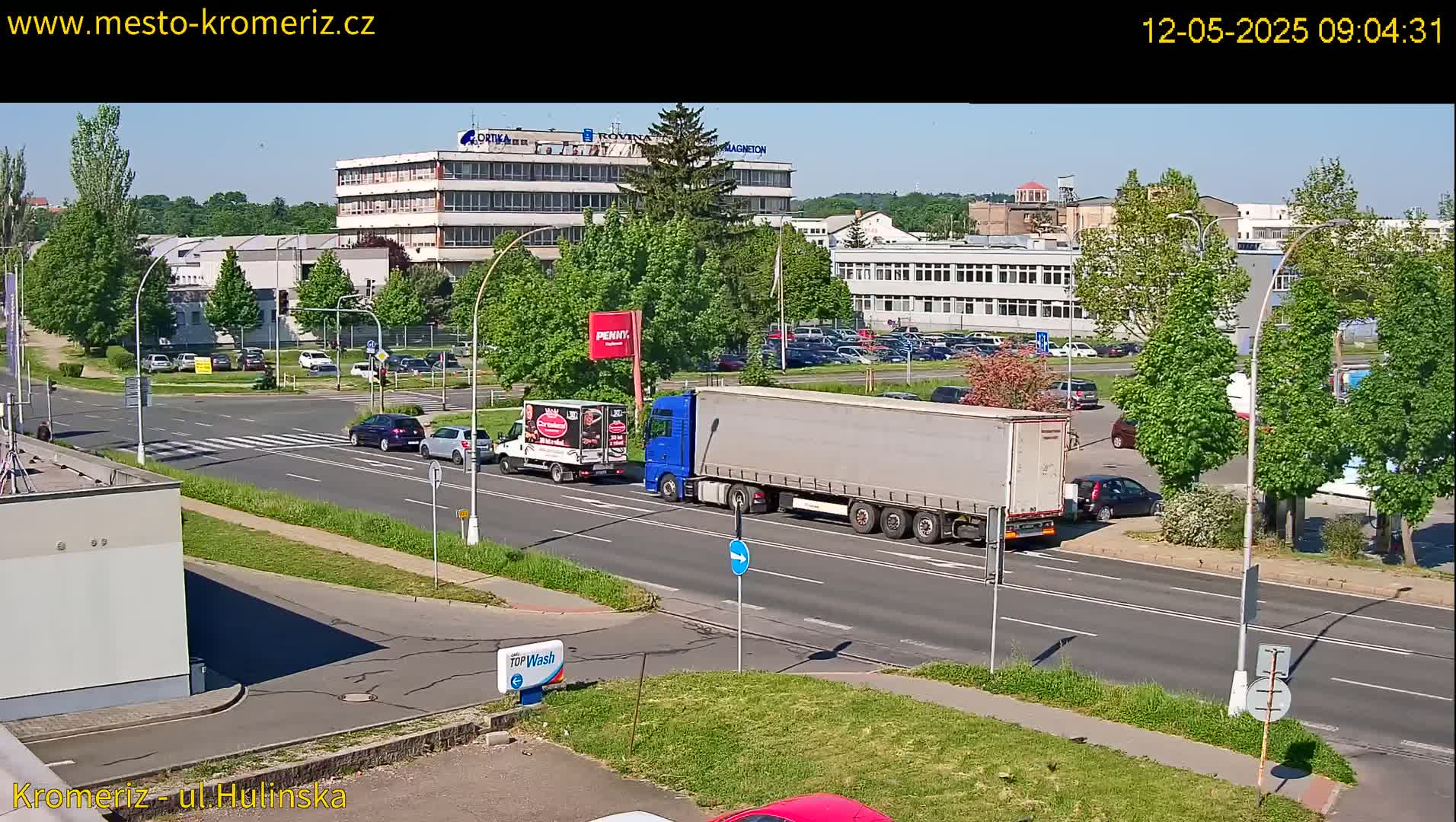 A sunny day reveals a multi-lane road with several cars and trucks passing an industrial area, surrounded by green trees and landscaping.