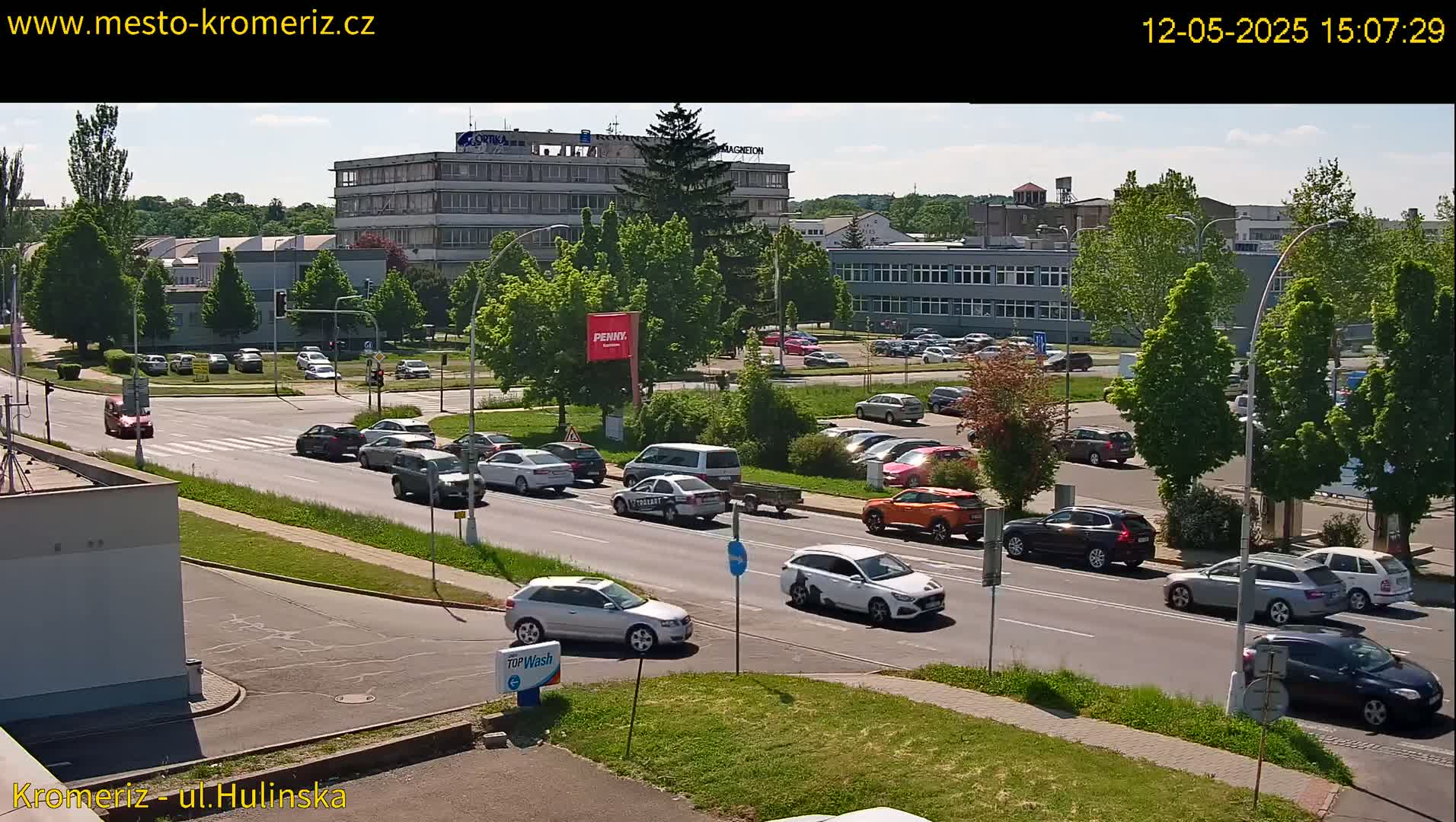 A sunny day reveals a multi-lane road with cars driving past industrial buildings and parking lots filled with vehicles, surrounded by lush green trees.