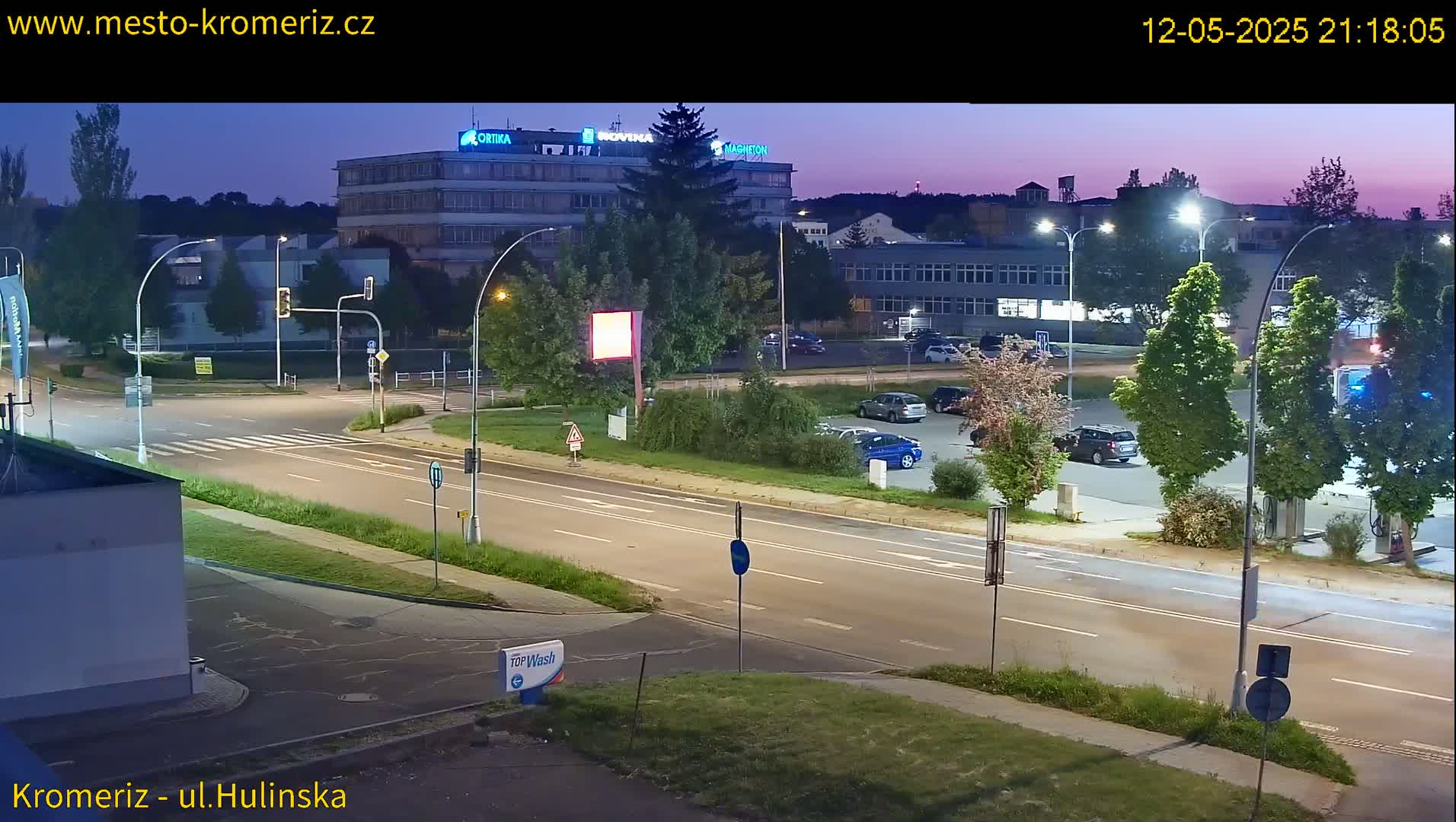 A twilight view of a multi-lane road with parked cars and streetlights illuminating a parking lot beside a large building.