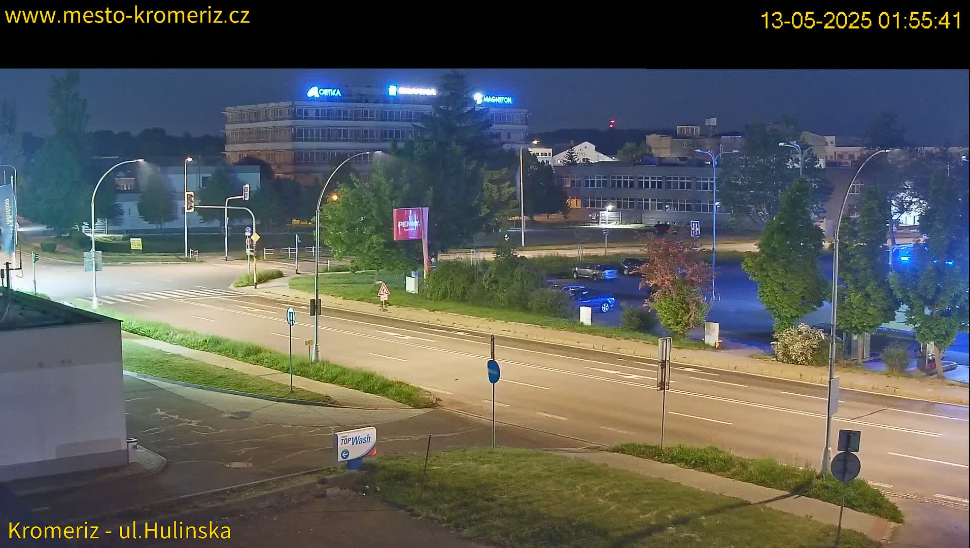A nighttime view of a multi-lane road with cars parked in a lot beside it, illuminated by streetlights, with several buildings visible in the background.