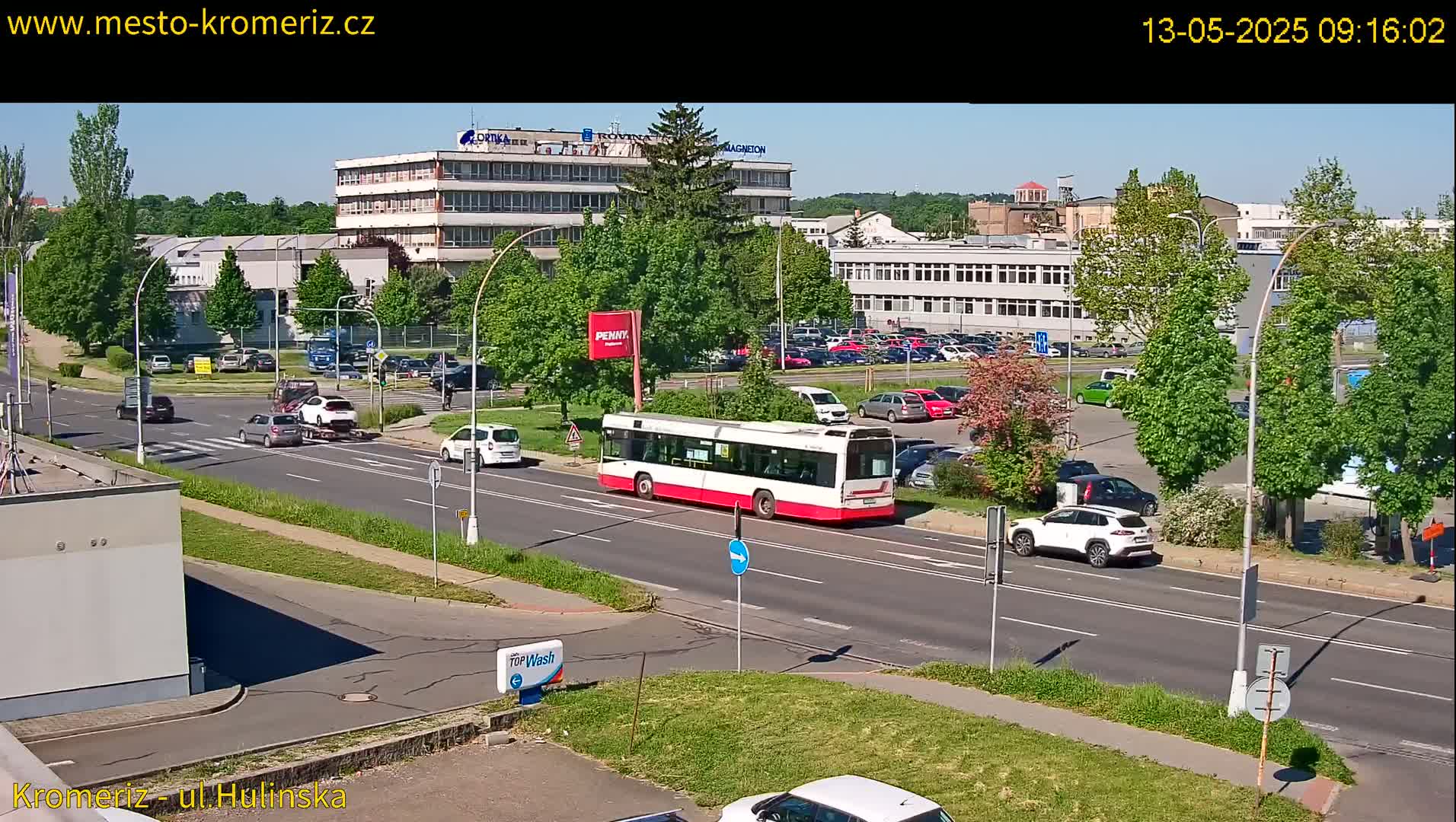 A sunny day with a red and white bus driving on a city street, surrounded by cars parked in lots and driving on the road, in front of several buildings and lush green trees.