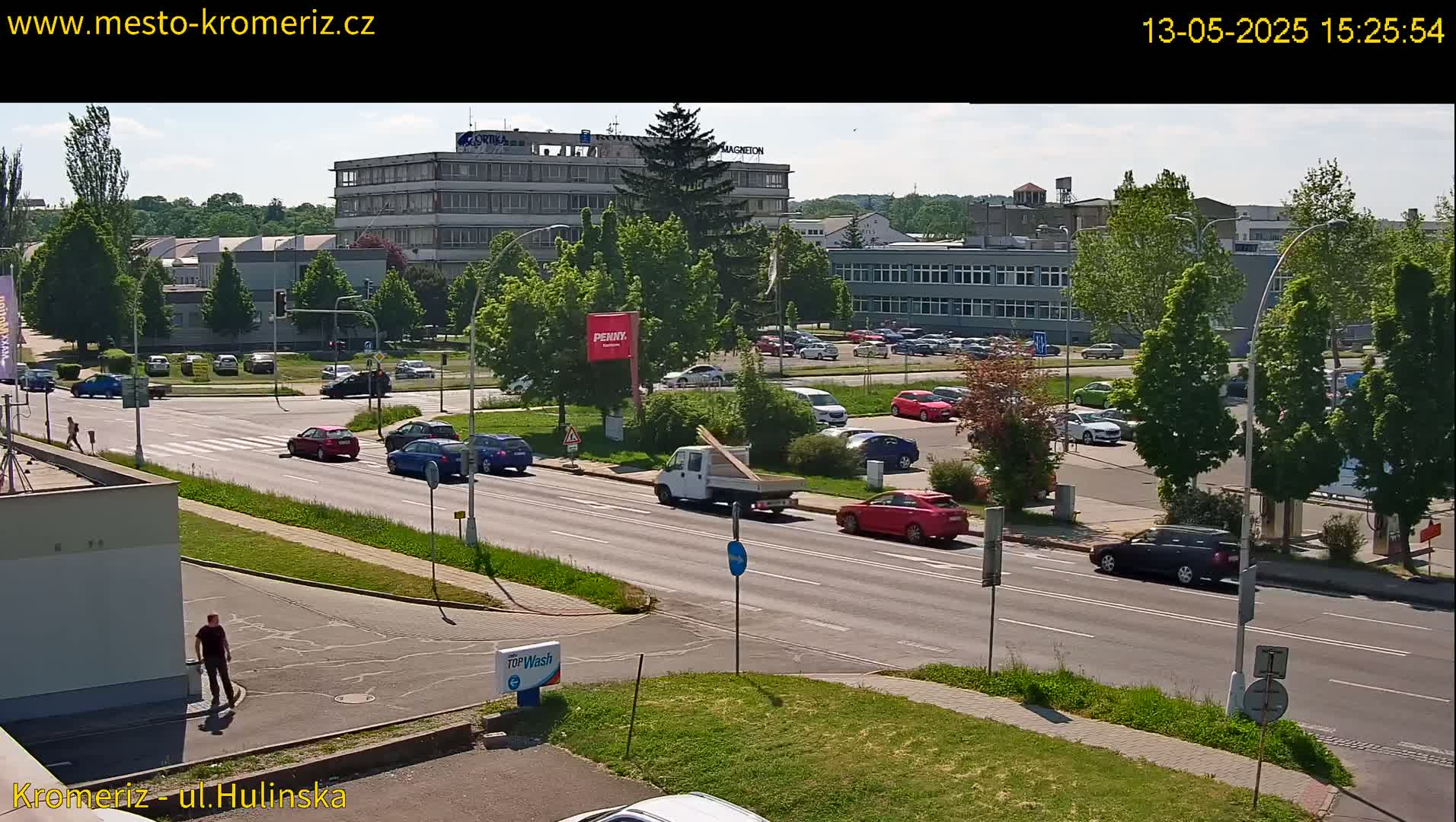 A sunny day shows a multi-lane road with cars driving past buildings and a parking lot filled with vehicles, surrounded by green trees and grass.