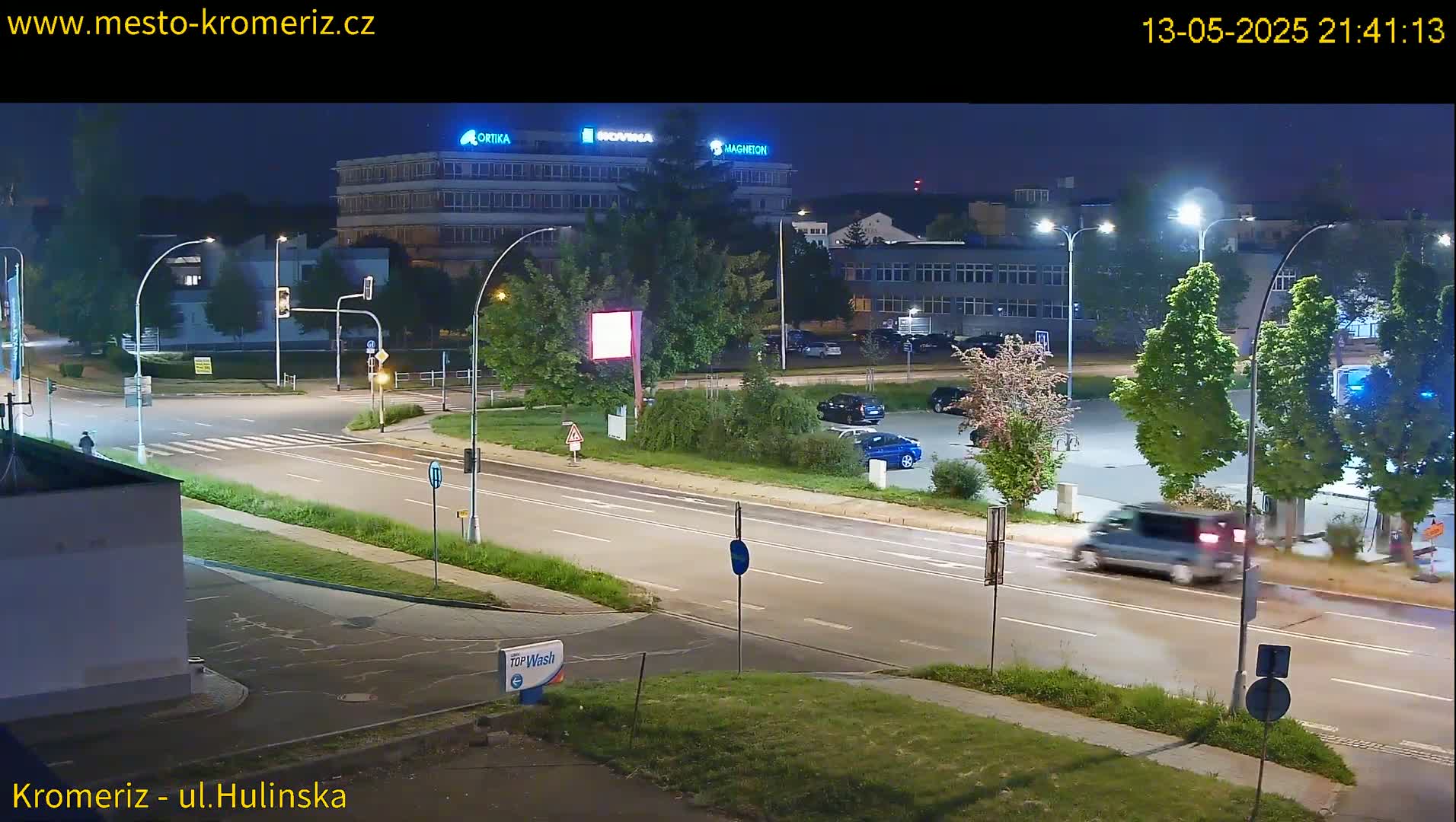 A nighttime street scene shows a multi-lane road with several cars and a van, under bright streetlights, near a large building and a parking lot with trees.