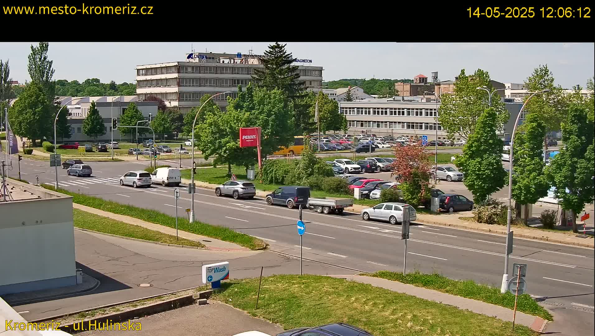A sunny day reveals a multi-lane road with several cars driving past office buildings and a parking lot filled with vehicles, all surrounded by lush green trees.