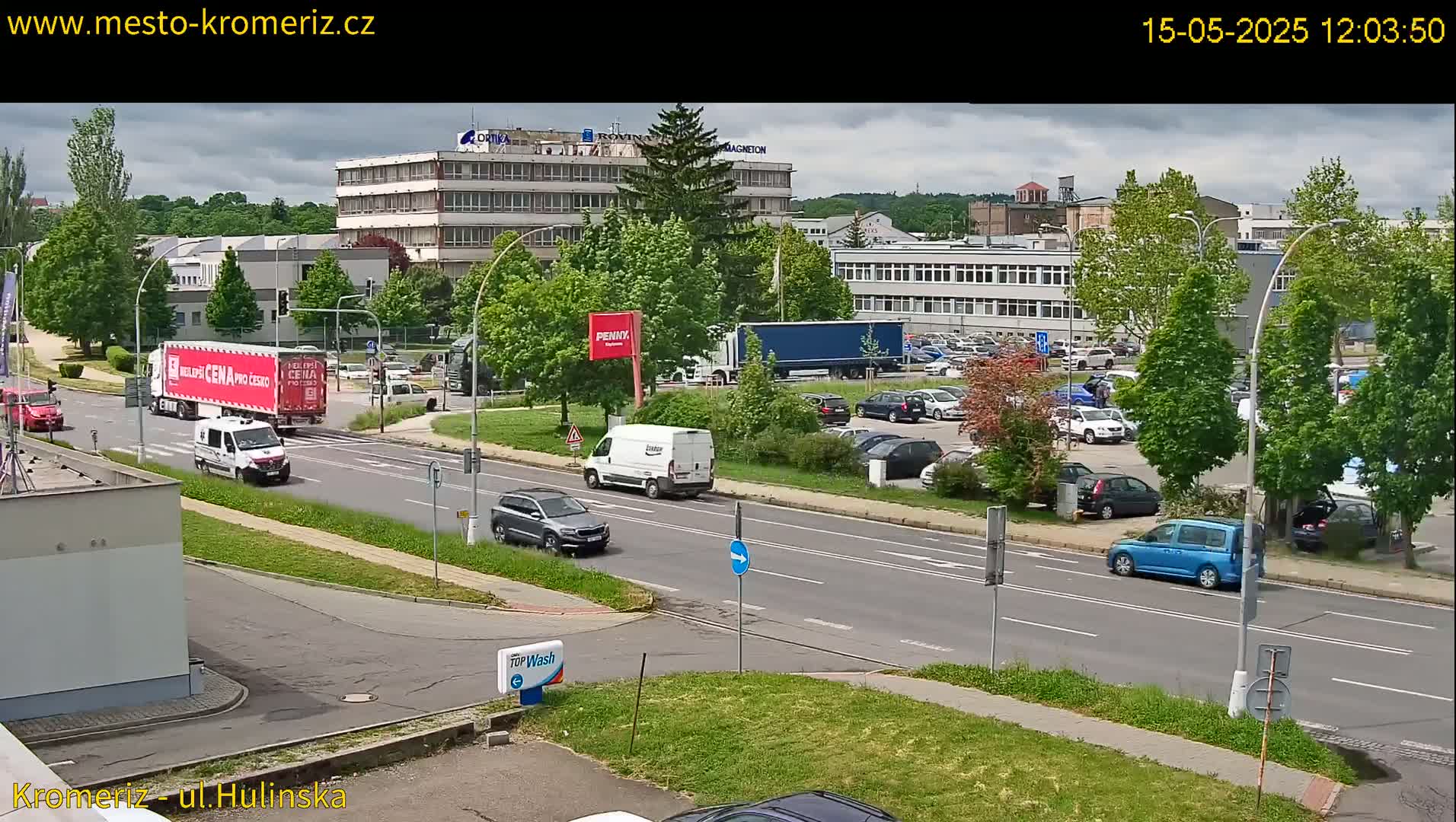 A partly cloudy day shows several vehicles driving on a road next to a parking lot and industrial buildings surrounded by green trees.
