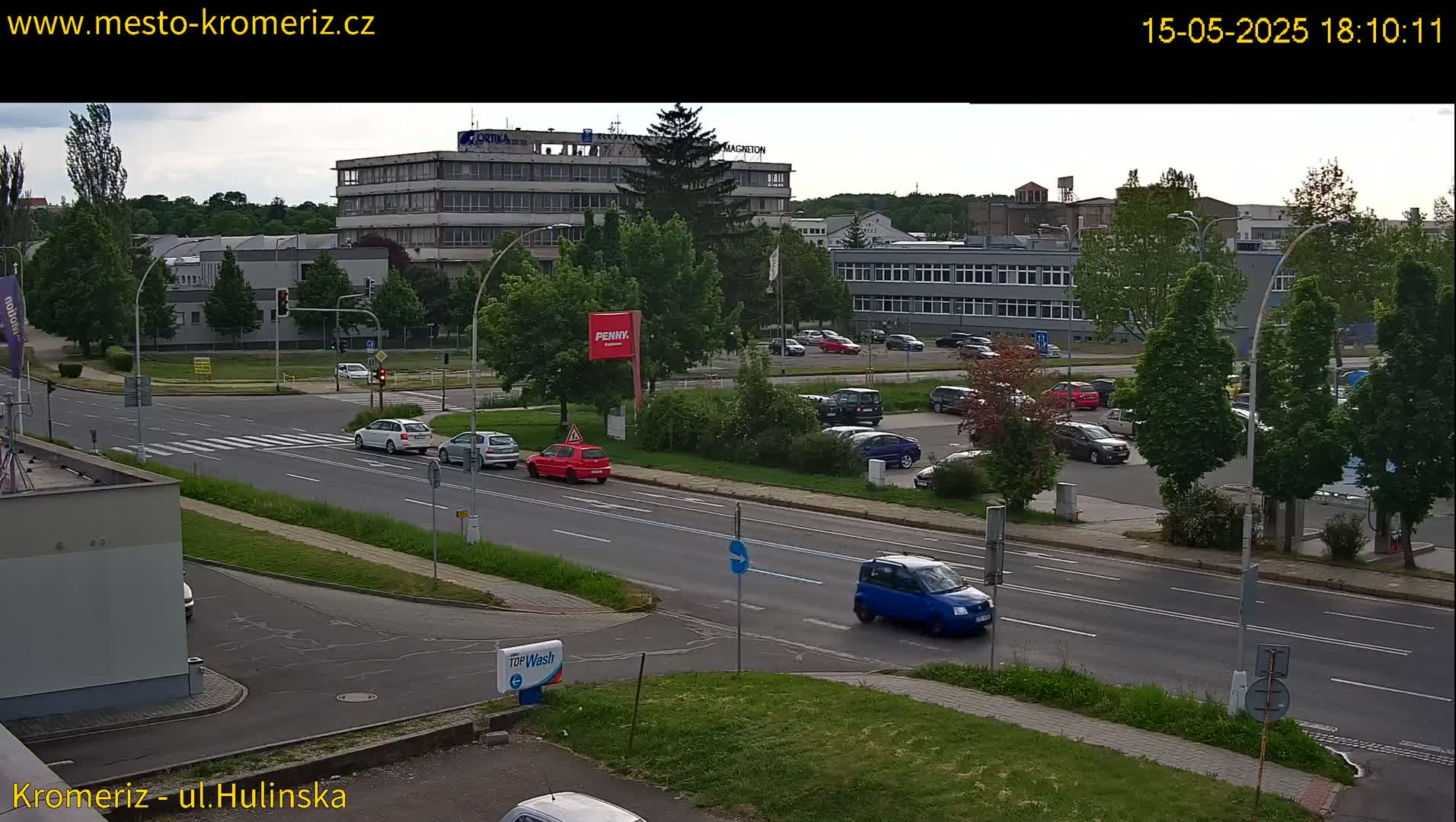A slightly overcast view of a multi-lane road with several cars driving and parked, near several multi-story buildings and lush green trees.