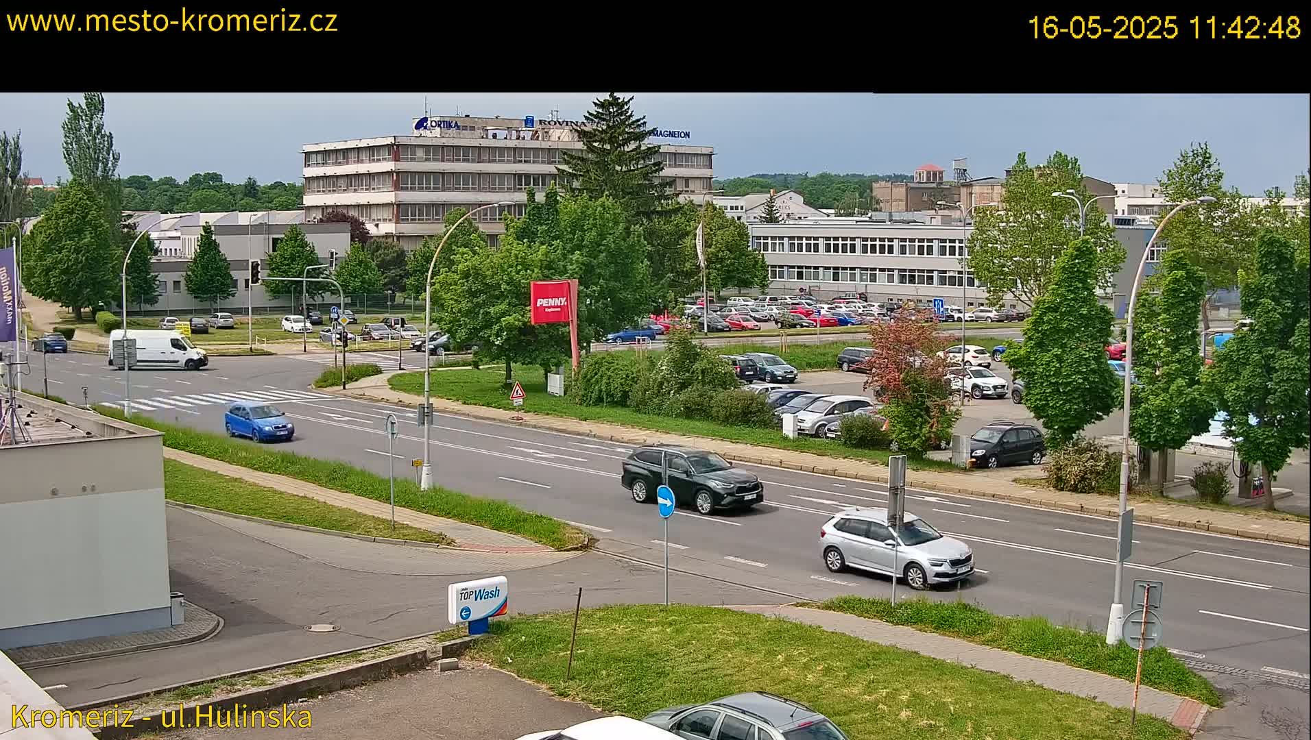 A sunny day reveals a multi-lane road with several cars driving past industrial buildings and a large parking lot filled with vehicles, all surrounded by lush green trees and landscaping.
