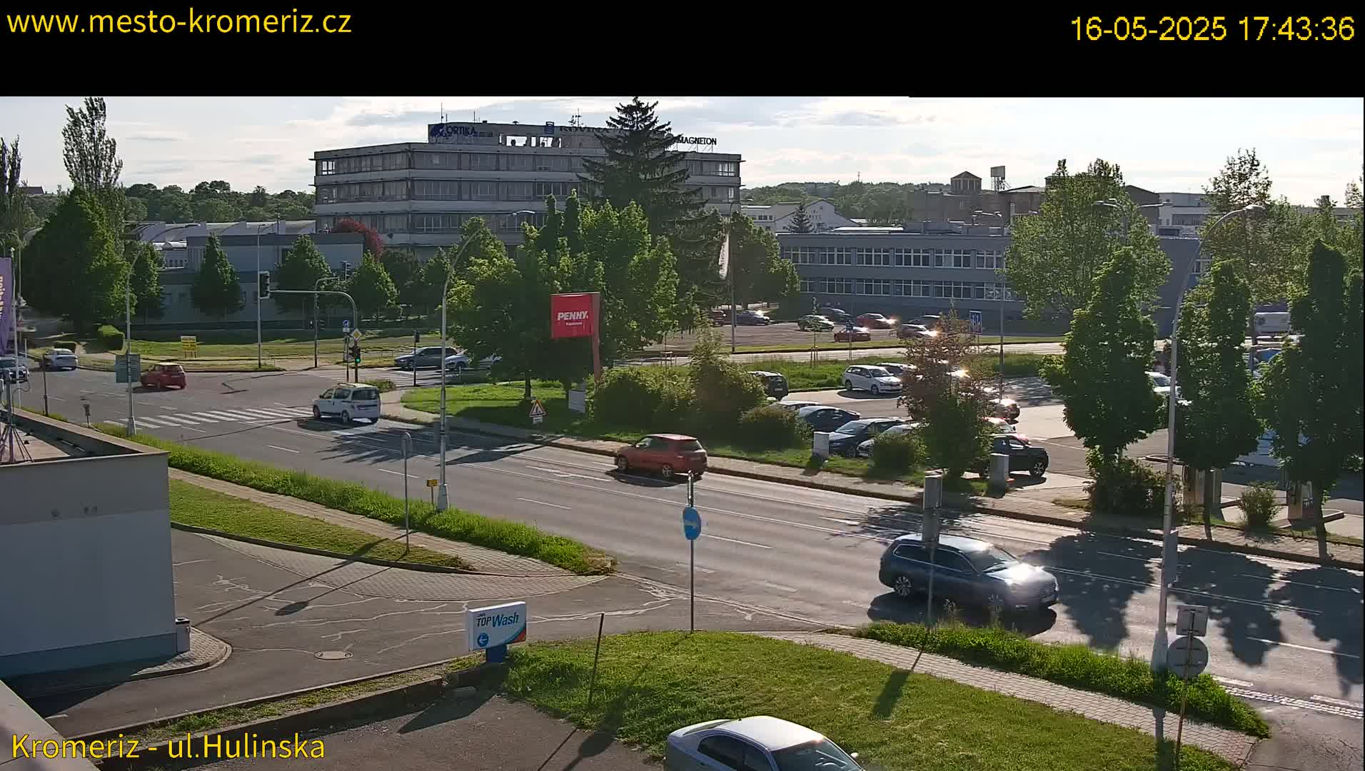 A sunny day shows several cars driving on a road that passes by a large building and parking lot with numerous other parked cars and lush green trees.