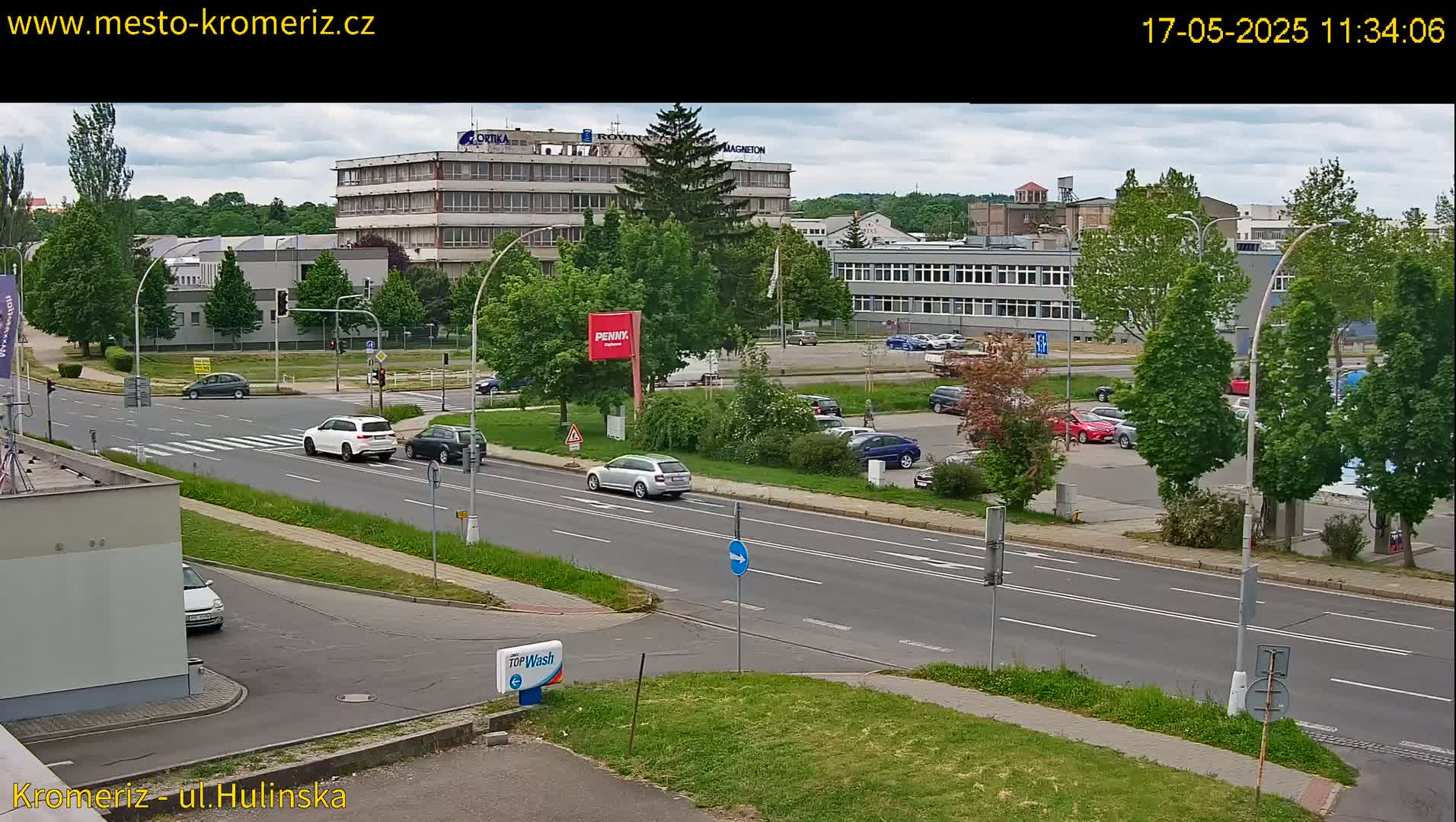 A partly cloudy day shows a street scene with several cars at an intersection near some buildings and a parking lot with lush green trees.