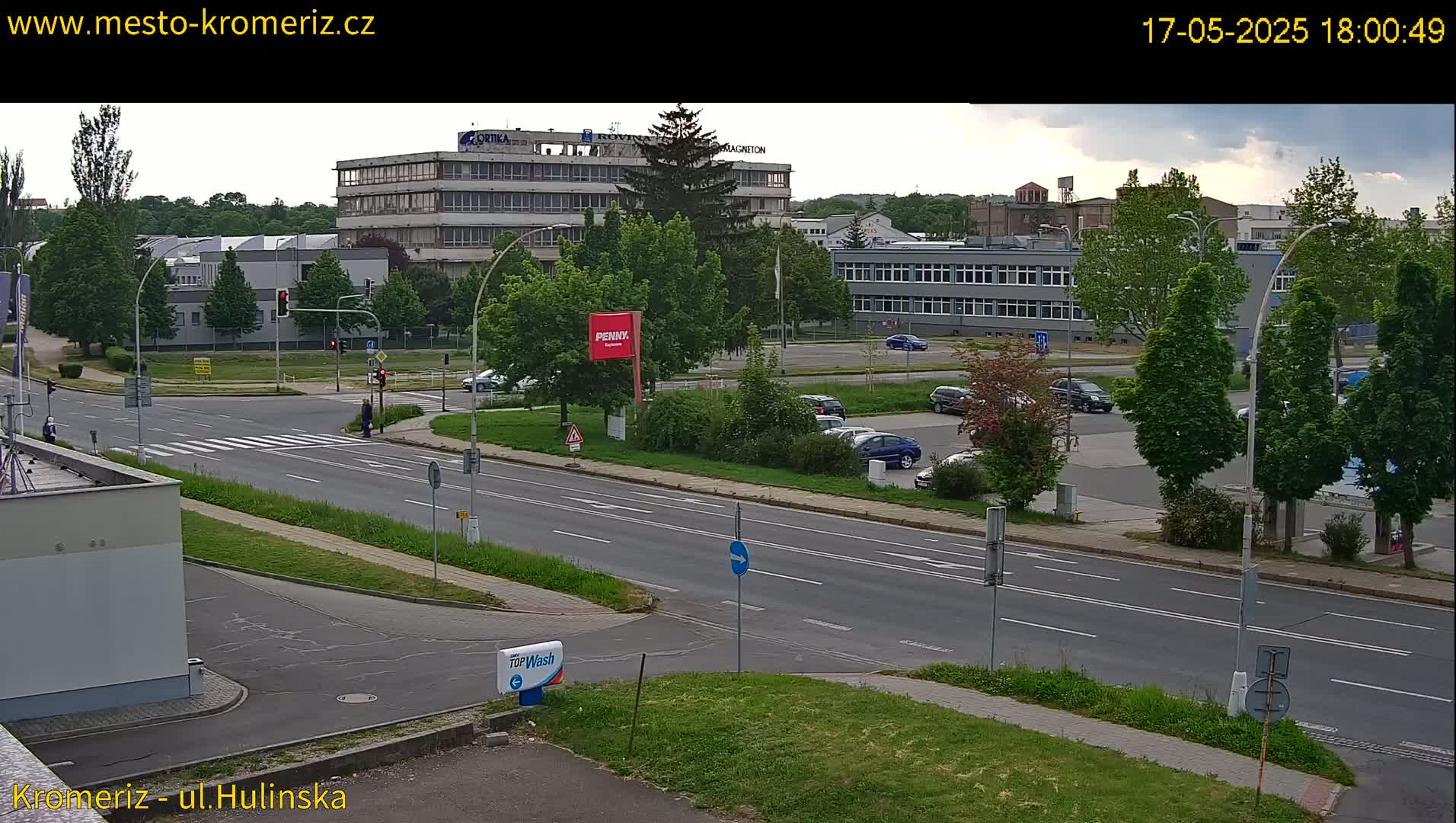 A mostly sunny street scene shows a multi-lane road intersecting with a crosswalk, with several cars parked in a lot and buildings in the background.