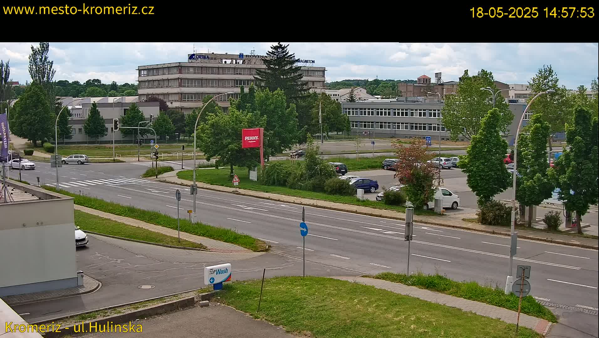 A partly cloudy day reveals a street scene with several cars parked in a lot next to a multi-story building and a roadway with cars driving.