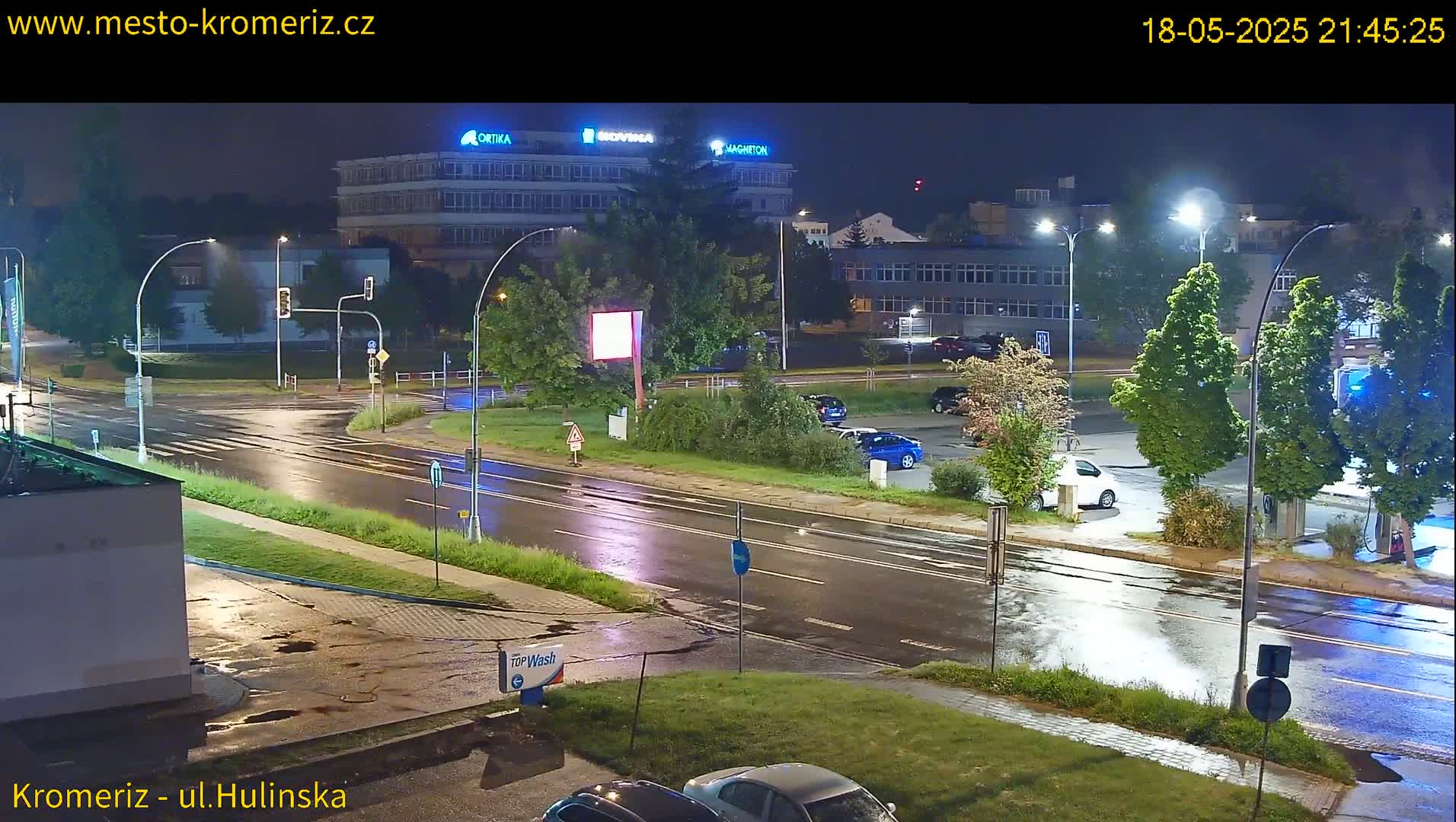 A nighttime view of a rain-slicked intersection with several cars parked nearby and buildings in the background under streetlights.