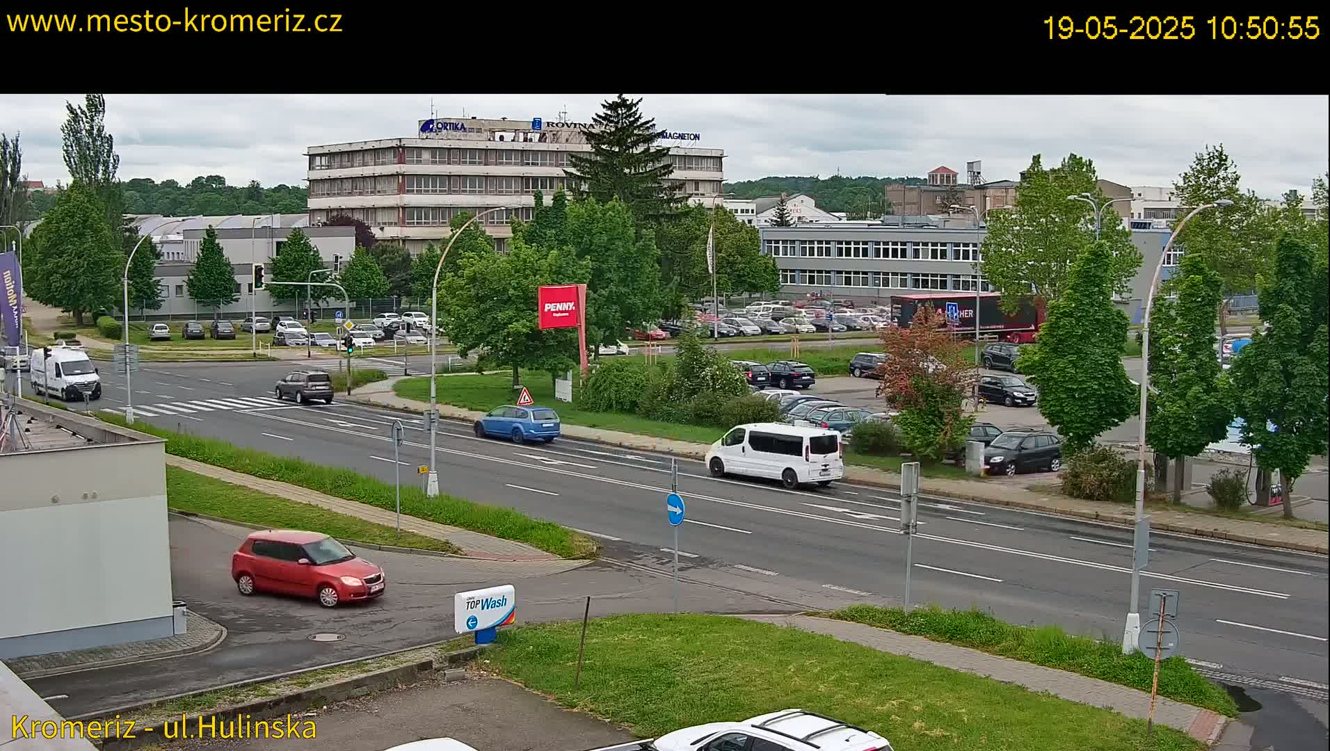 A mostly sunny day shows several cars driving on a multi-lane road near industrial buildings and a parking lot with many cars, with lush green trees and grass along the road and in the background.