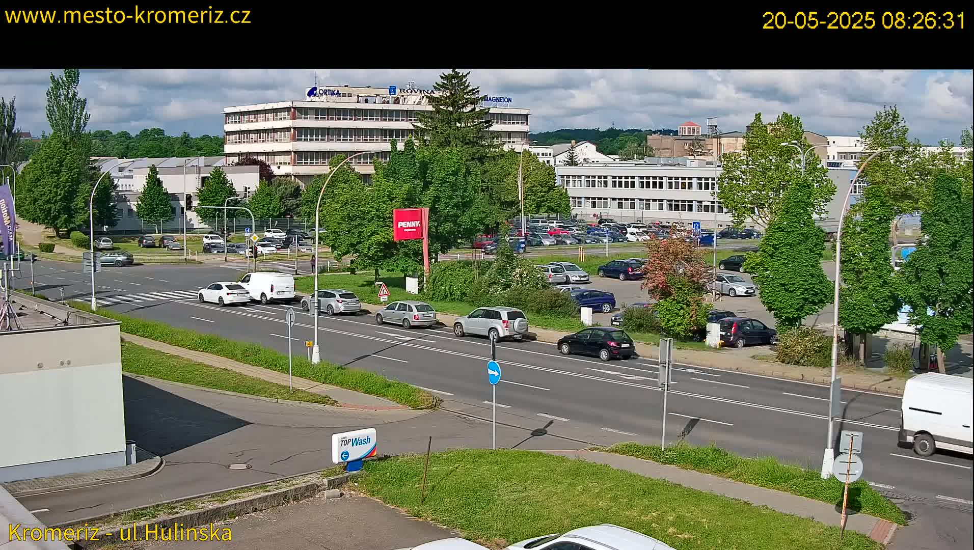 A sunny day shows a street scene with several cars driving on a multi-lane road, passing by a large parking lot and several multi-story buildings.