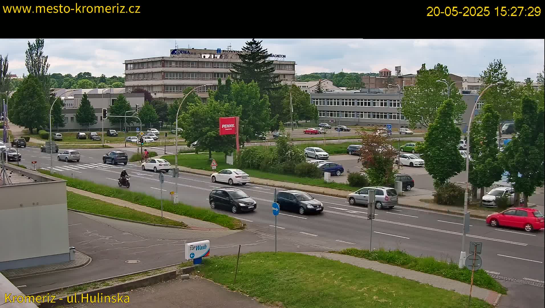 A mostly sunny day reveals a street scene with multiple cars and a motor scooter traversing a multi-lane road that intersects with a smaller road, all in front of several buildings and a parking lot filled with vehicles.