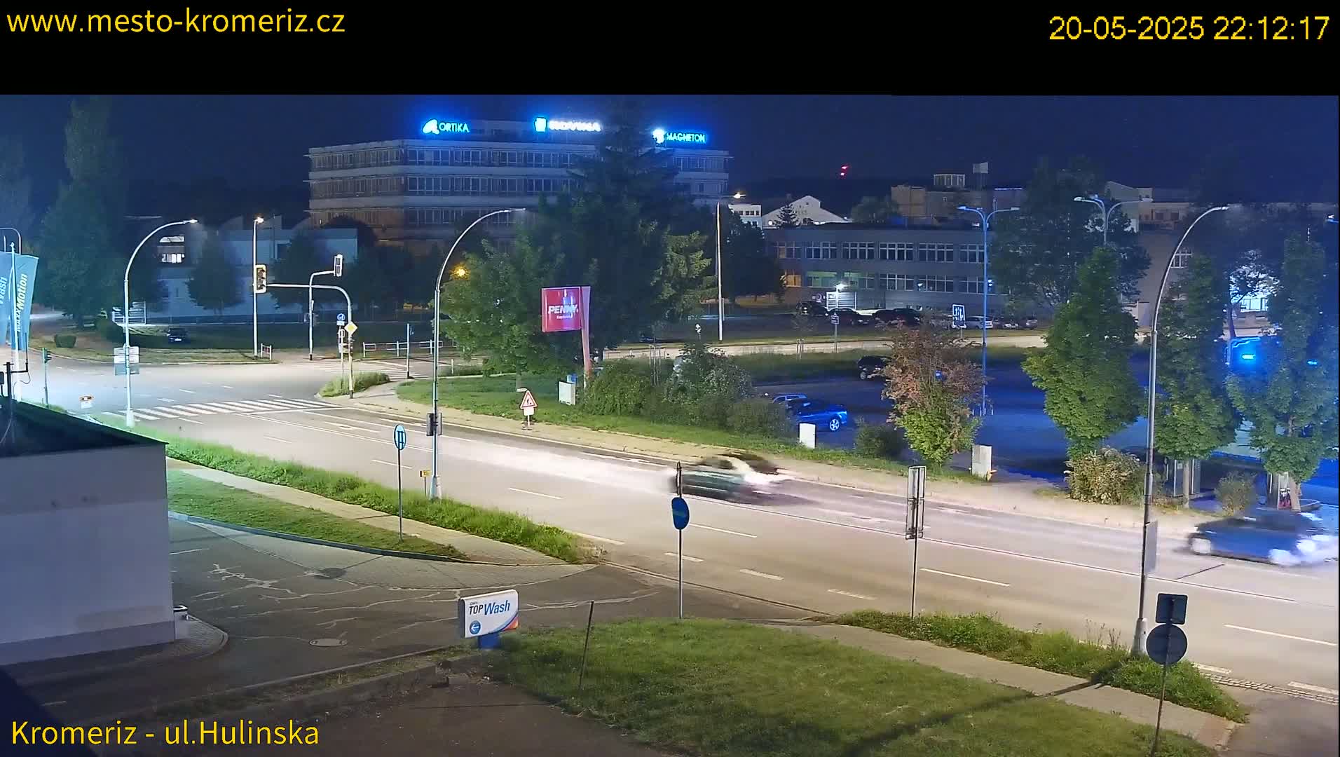A nighttime view of a multi-lane road with cars driving past buildings and streetlights under a clear sky.