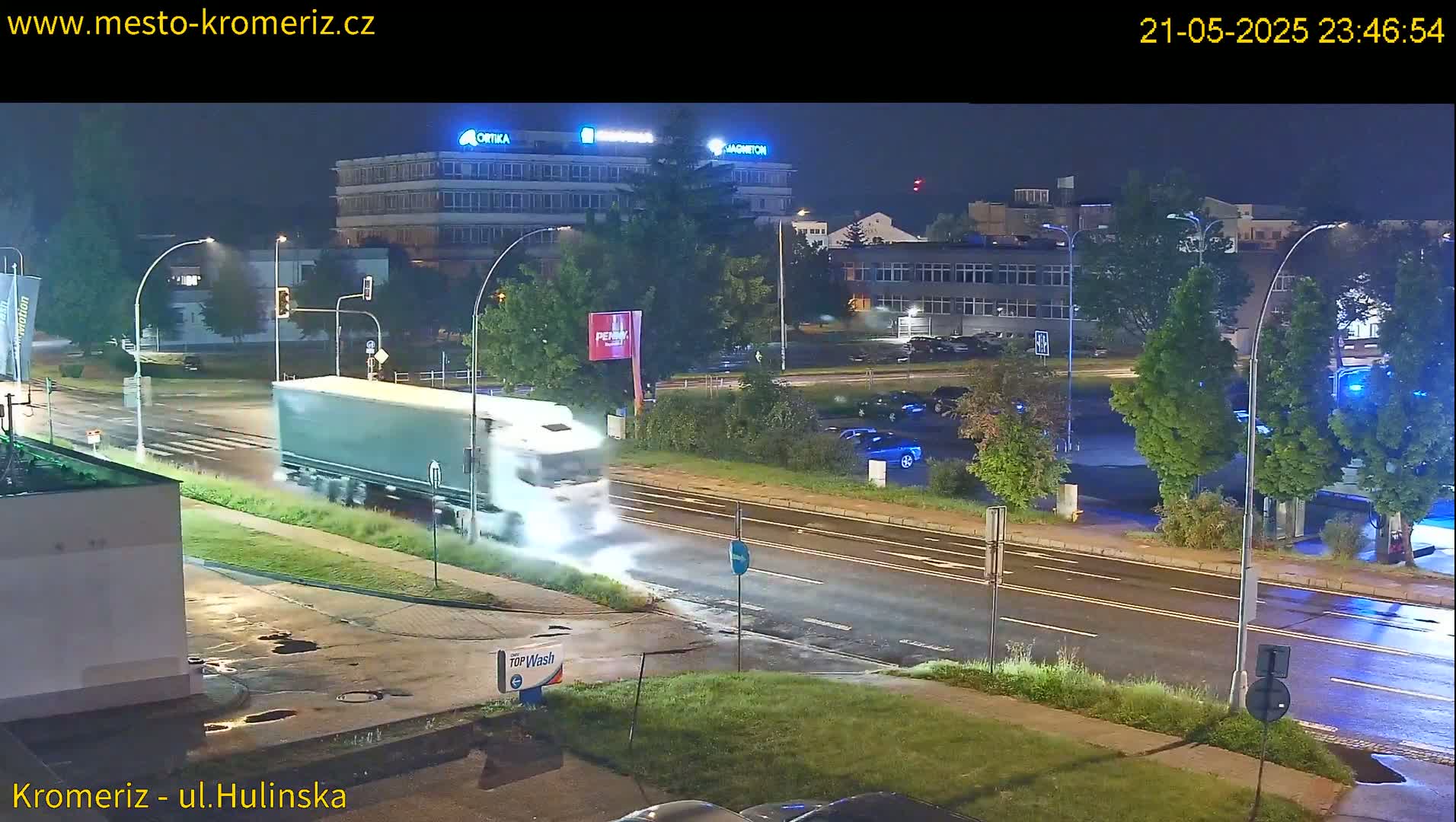 A large truck is driving on a wet, nighttime street past several buildings and parked cars.
