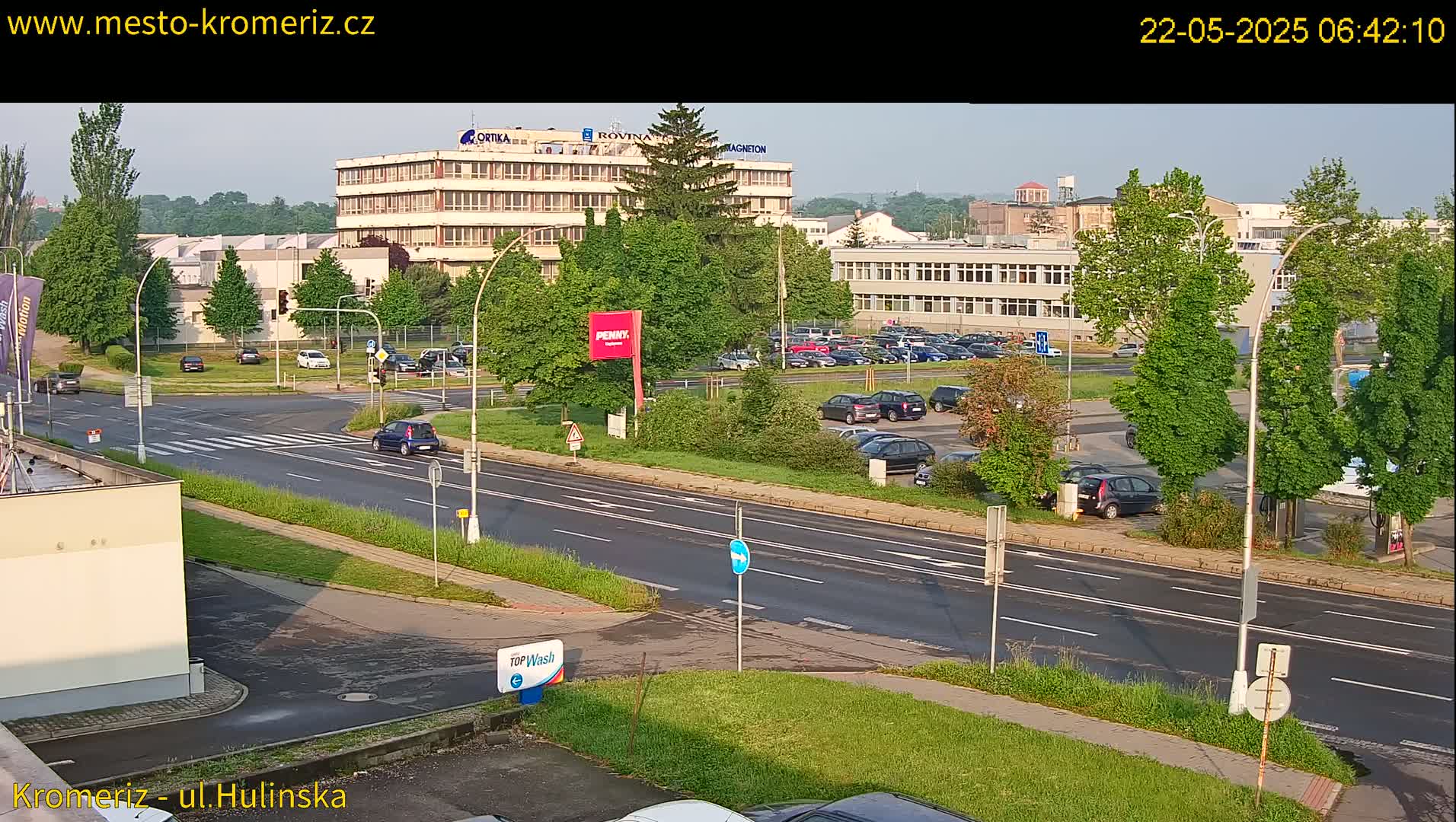 A sunny day reveals a multi-lane road passing by a parking lot full of cars and a large industrial building, with lush green trees lining the road and surrounding the area.