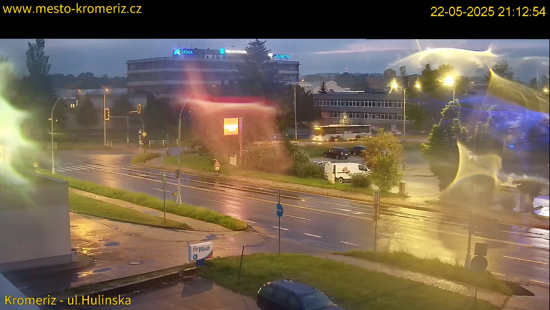 A wet road at night shows a few cars and a bus, with buildings and streetlights visible in the background under a partly cloudy sky.
