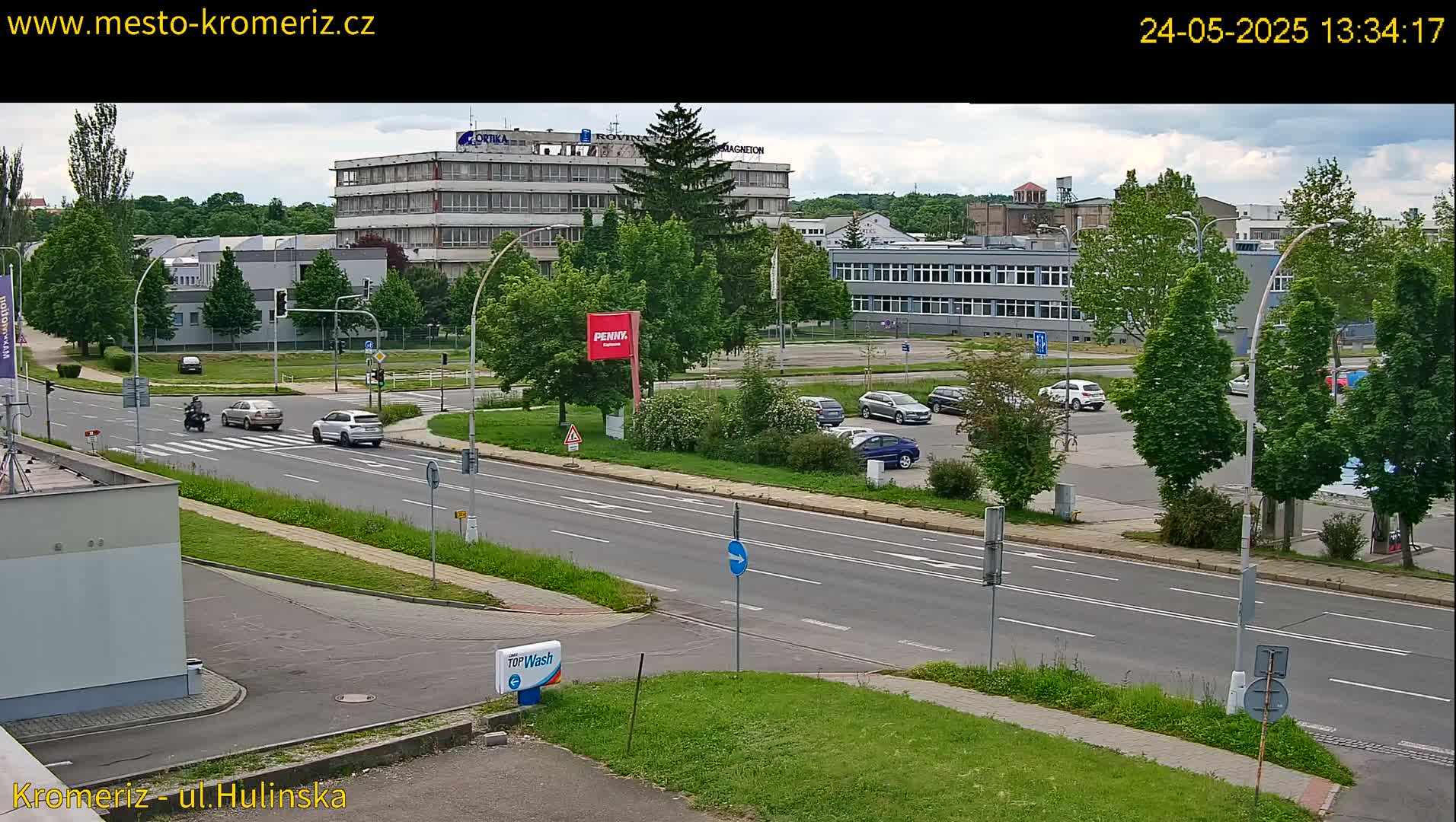 A partly cloudy day shows a multi-lane road with several cars and a motorcyclist, passing by industrial buildings and a parking lot with more vehicles.