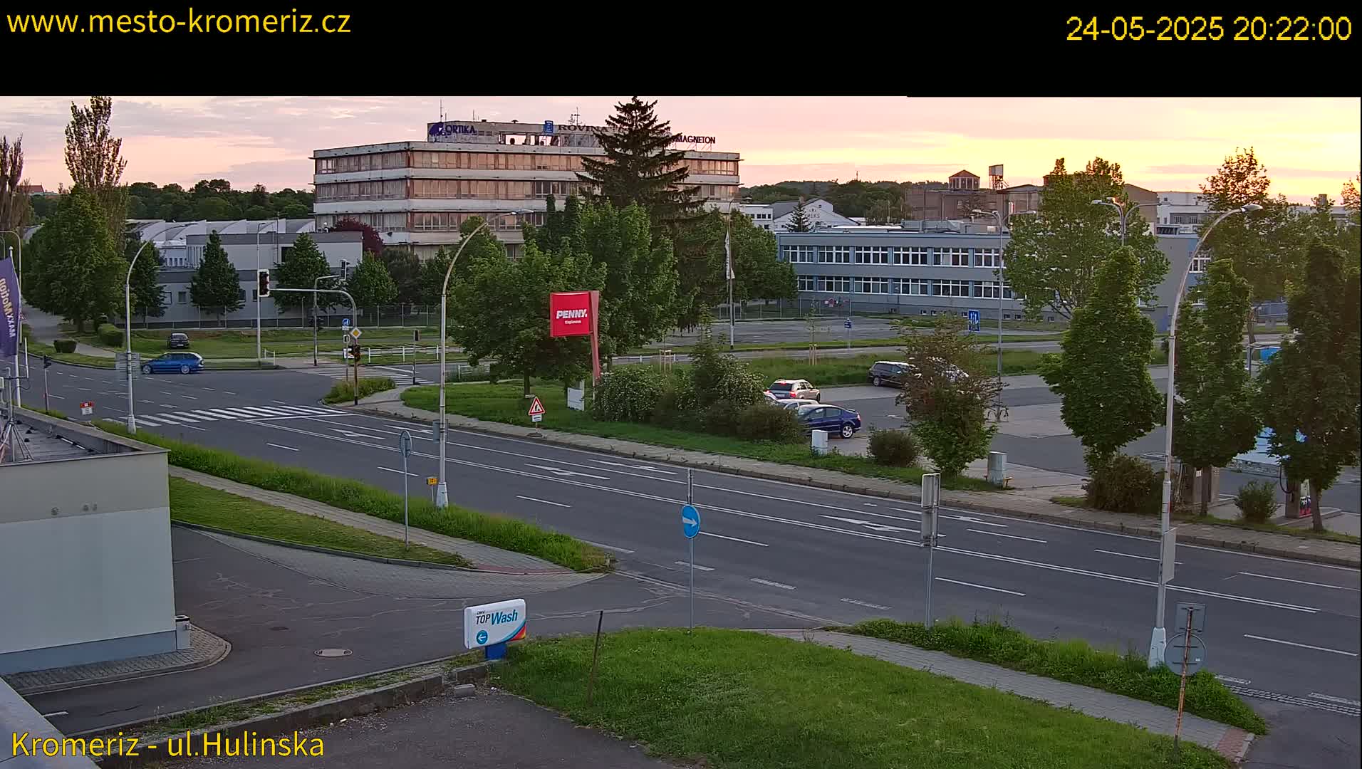 A wide shot of a road intersection at dusk, with several buildings and cars visible under a mostly clear, sunset sky.