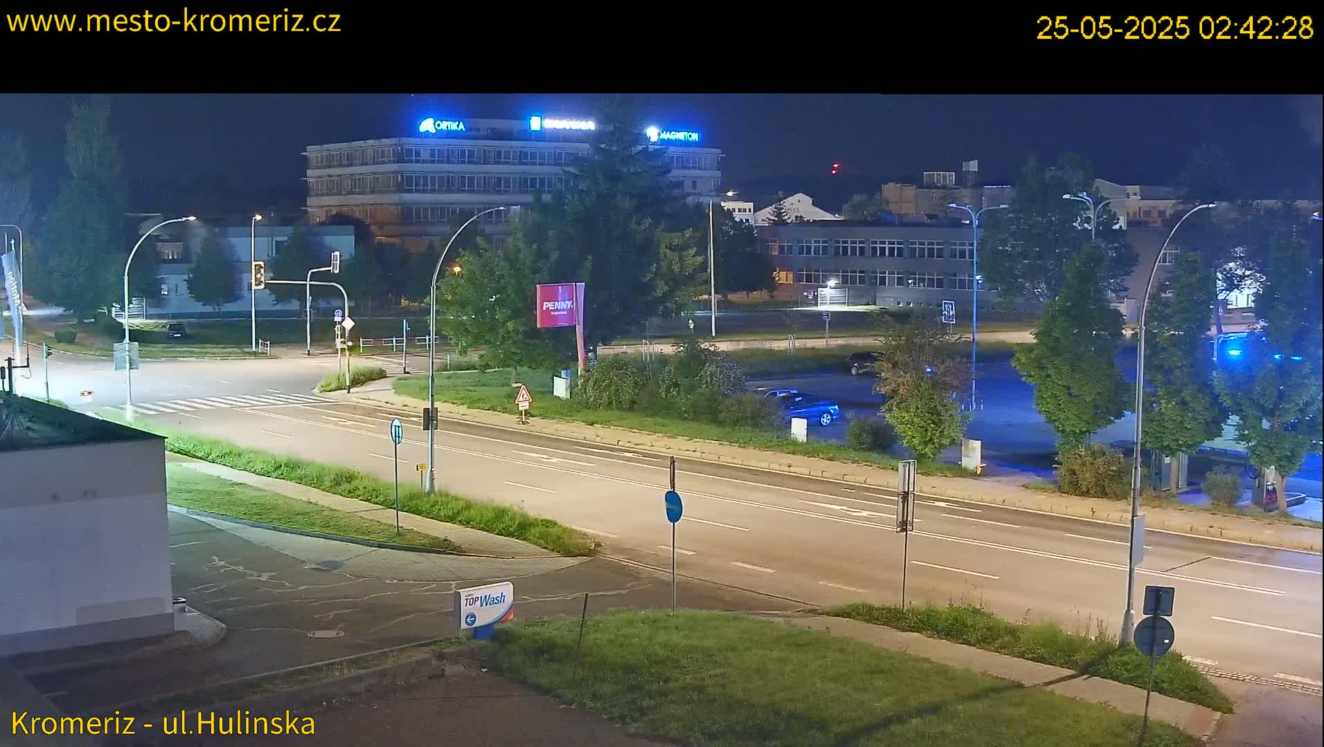 A nighttime, street-level view shows a multi-lane road intersecting with another road, surrounded by buildings, trees, and streetlights under a clear sky.