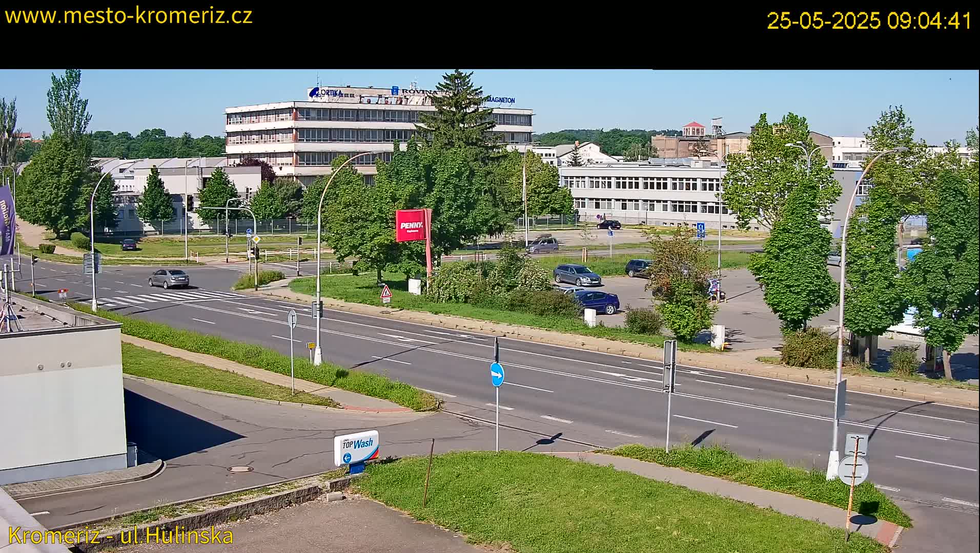 A sunny day reveals a multi-lane road with several cars, passing in front of industrial buildings and a parking lot surrounded by lush green trees.
