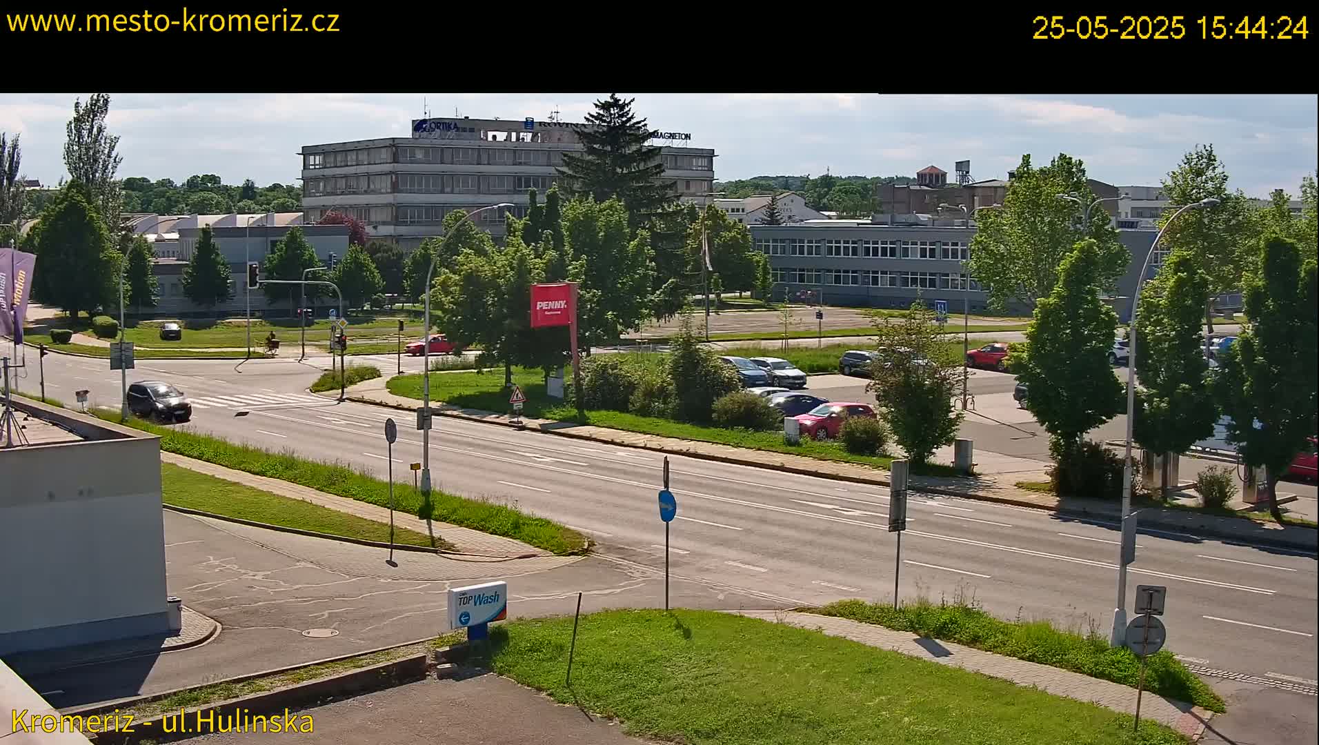 A sunny day view of a multi-lane road with cars and buildings, including a large multi-story building, and a parking lot with several parked cars.