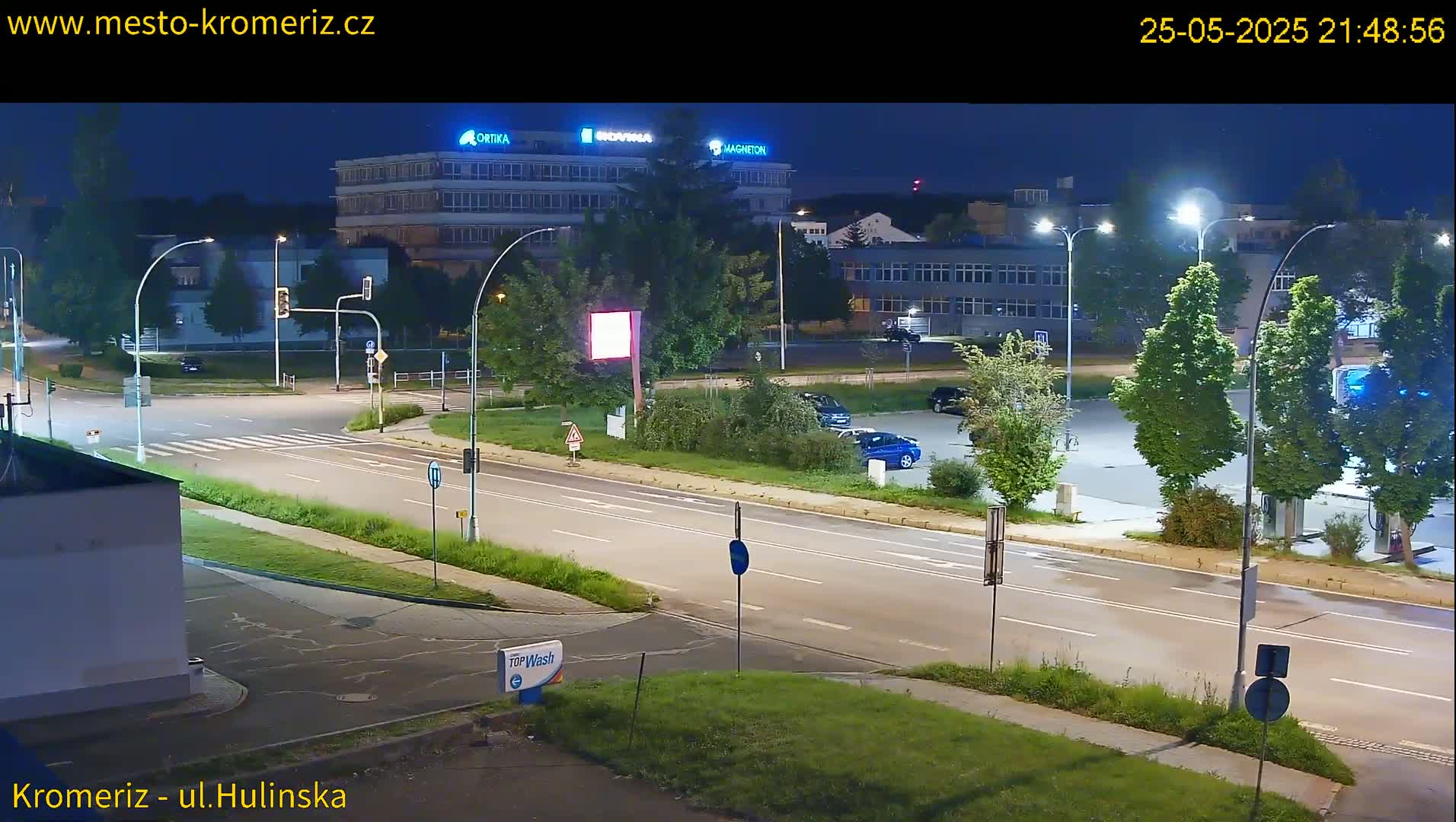 A nighttime street scene shows a multi-lane road with cars parked in a lot across from a large building, under bright streetlights.