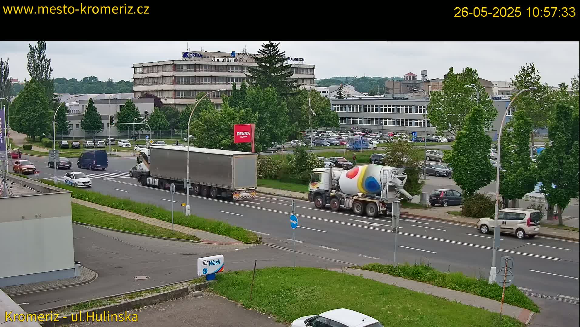 A cement mixer and several cars are driving on a city street under an overcast sky, with large buildings and lush green trees in the background.