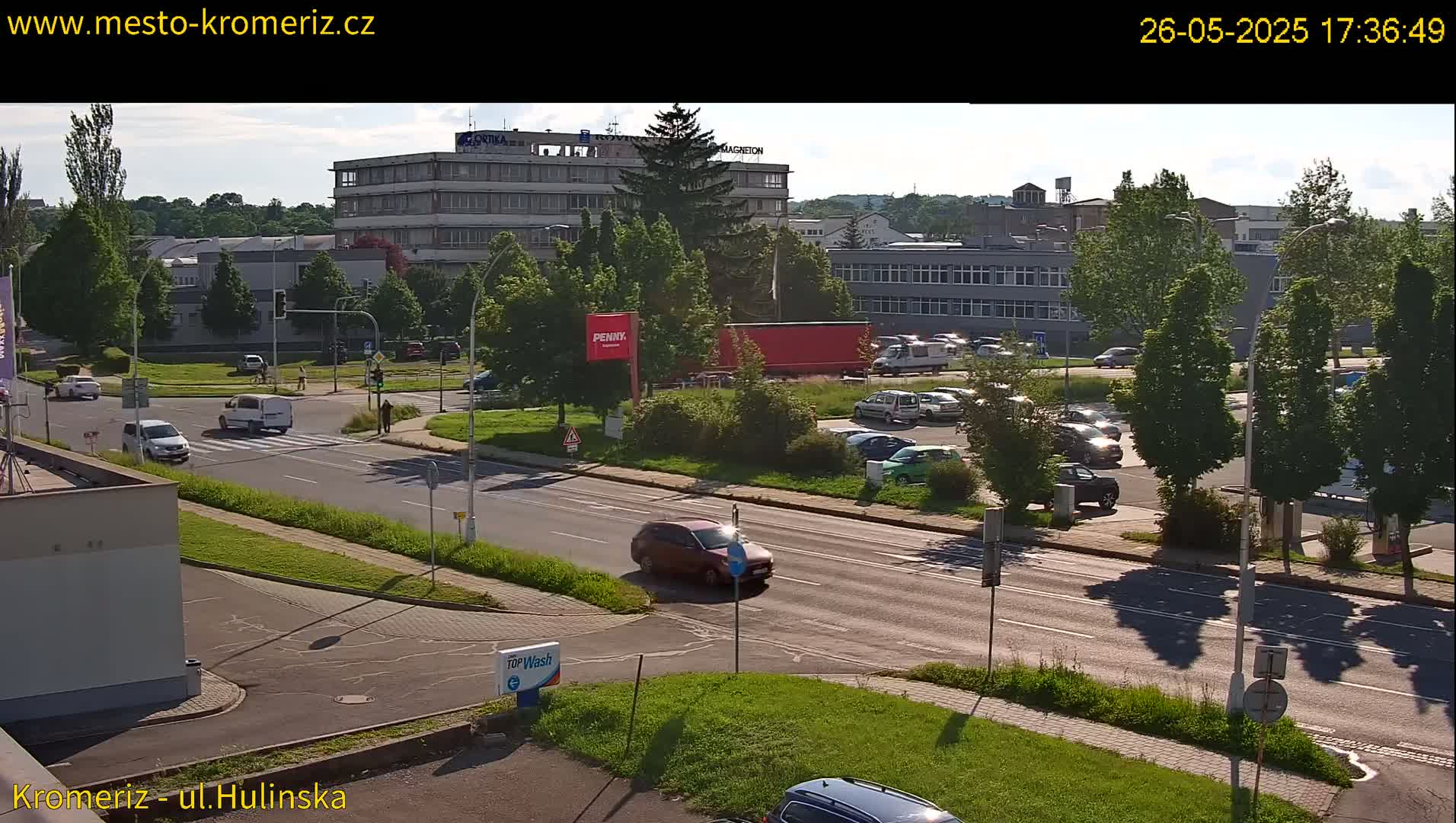 A sunny day reveals a street scene with several cars driving on a multi-lane road, passing a parking lot filled with vehicles near buildings and green trees.