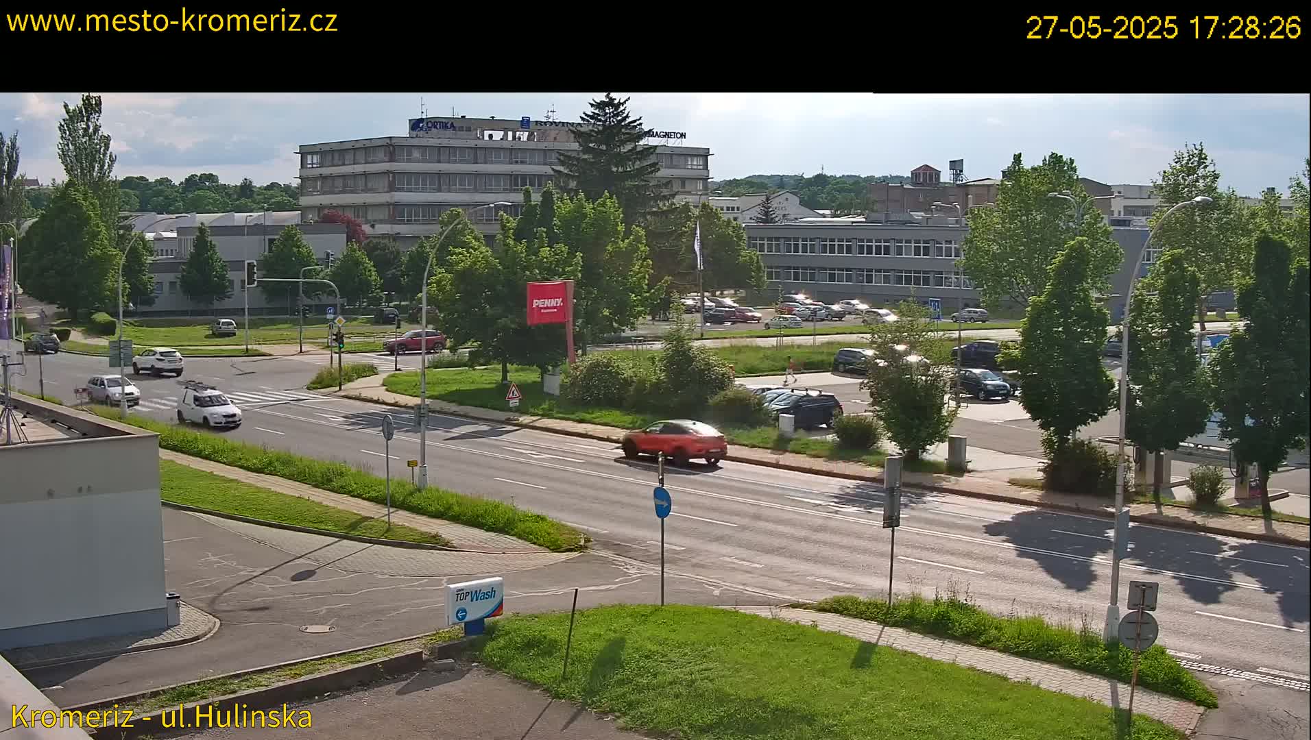 A sunny day shows a road intersection with several cars and a parking lot next to buildings, including a multi-story office building and other structures.