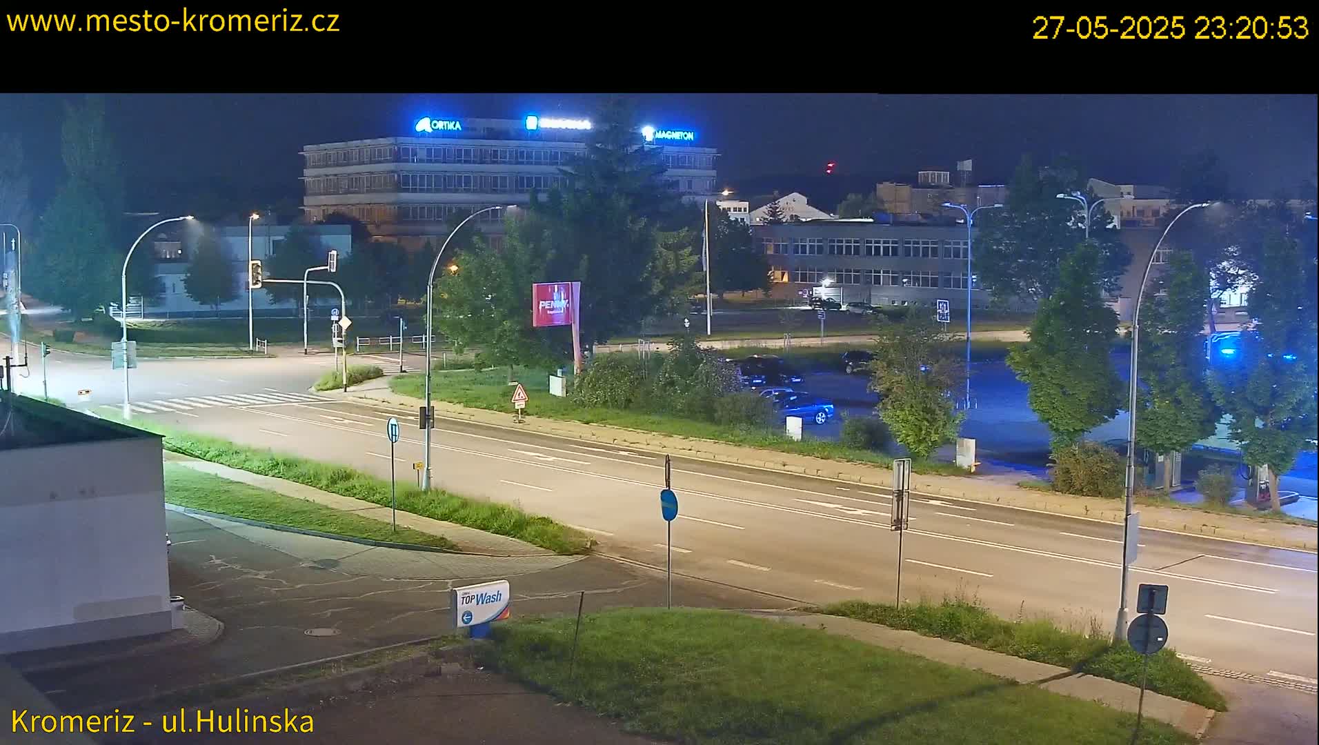 A nighttime view of a mostly empty road and intersection, with several buildings and parked cars visible in the background under partly cloudy skies.