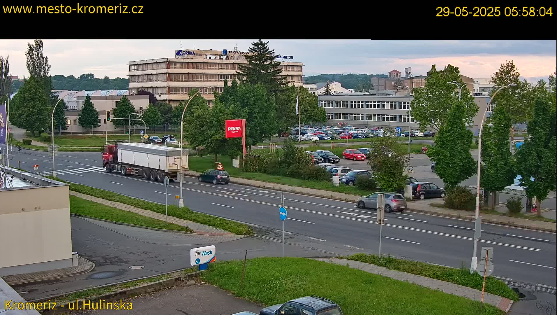 A partly cloudy morning scene shows a large semi-truck and several cars driving on a multi-lane road in front of industrial buildings and a parking lot filled with vehicles.