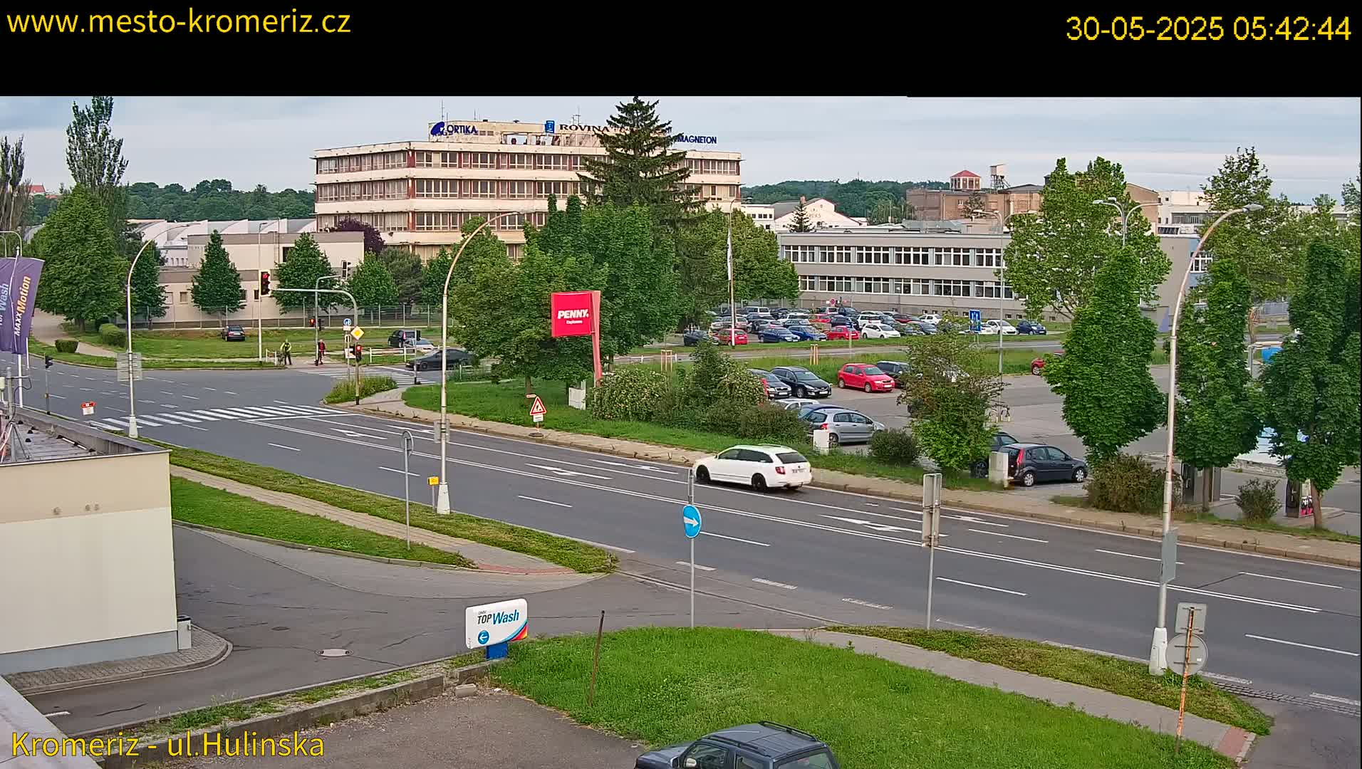 A mostly sunny, daytime view of a multi-lane road in front of several industrial buildings and a large parking lot filled with cars, with several pedestrians crossing the street at a crosswalk.