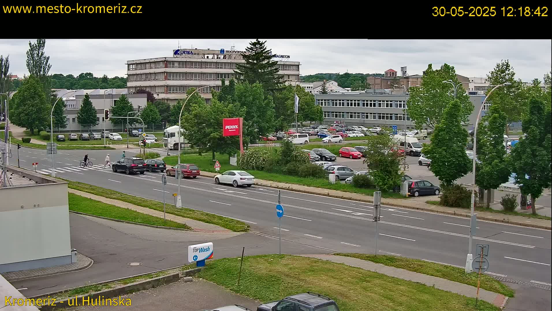 A mostly sunny day shows several cars driving on a multi-lane road that passes in front of several buildings and a large parking lot filled with cars.