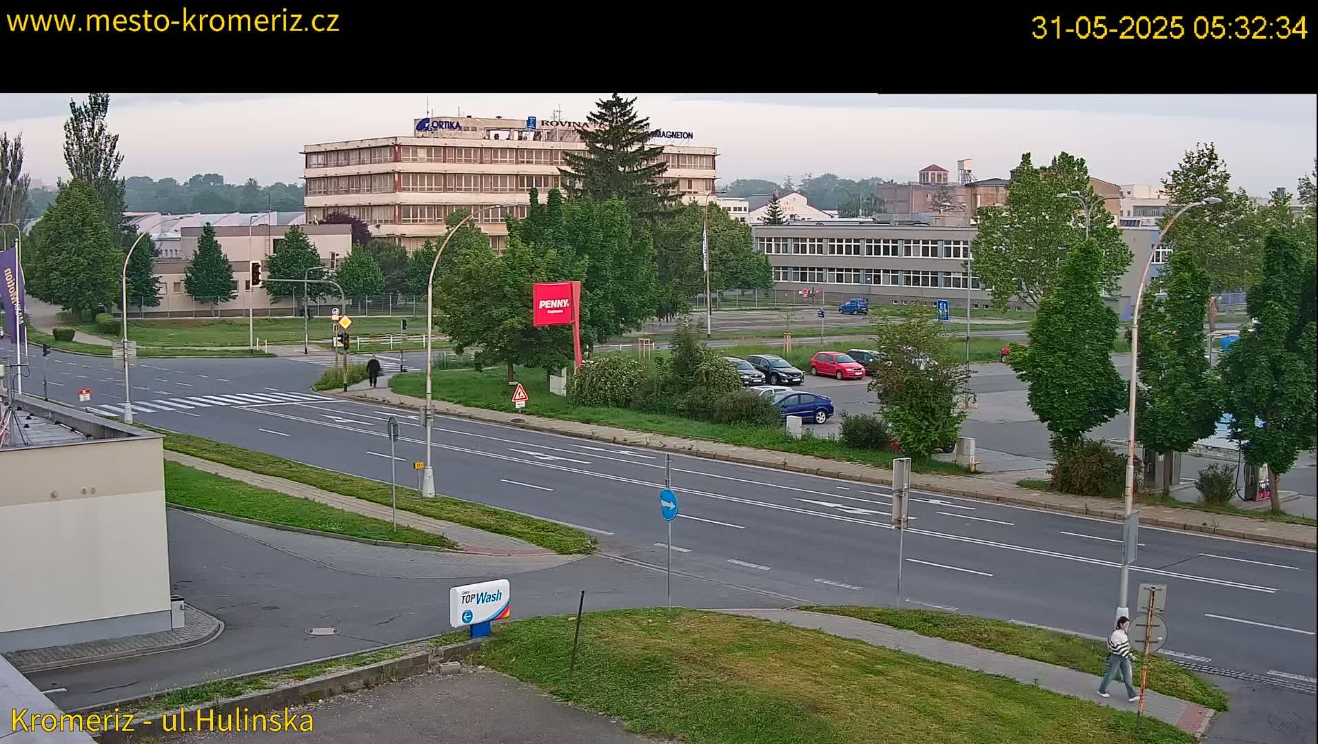A street scene on a partly cloudy morning shows a multi-lane road, several cars parked in a lot, and several buildings including what appears to be a factory or industrial complex, with pedestrians walking on sidewalks.