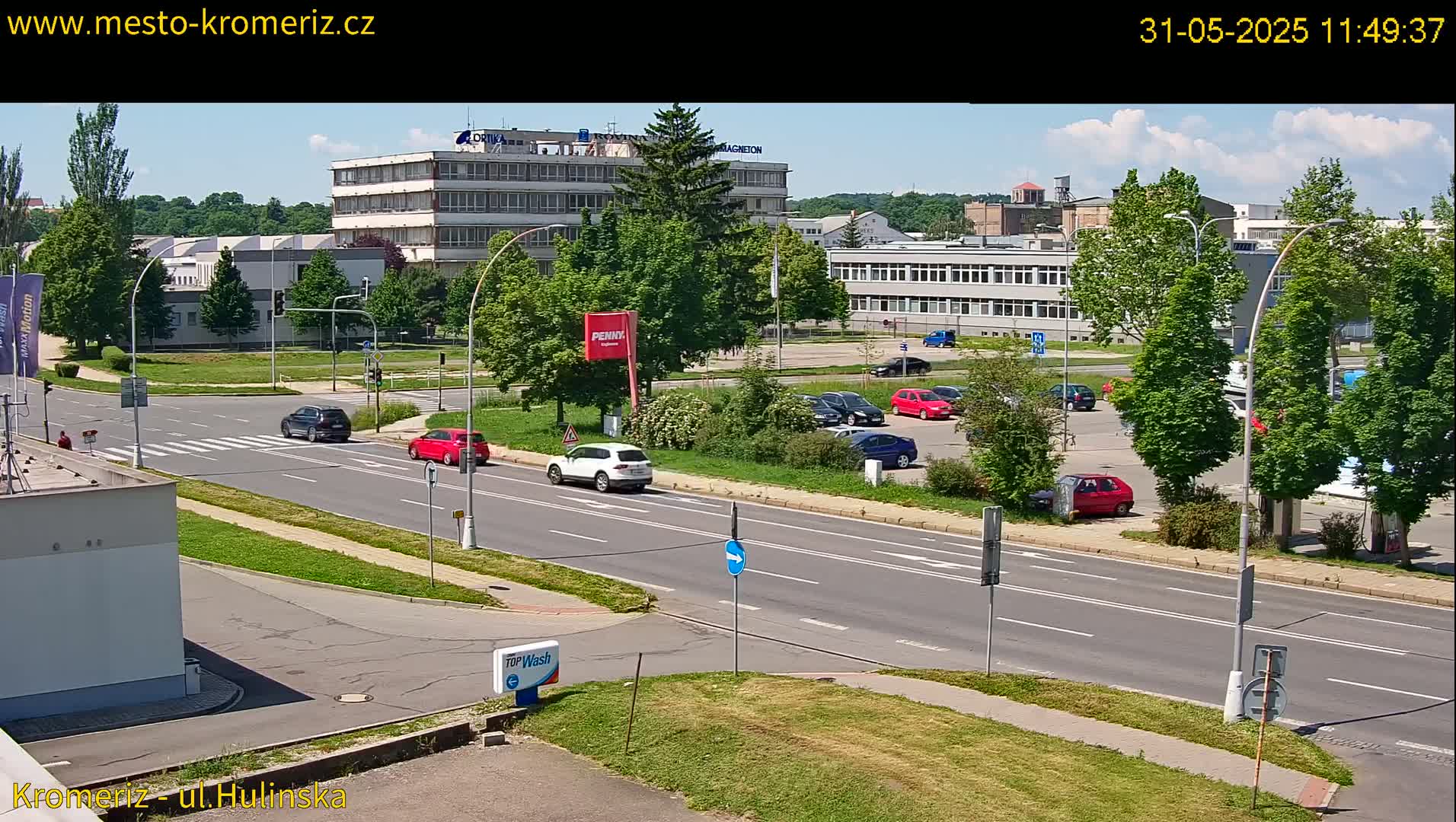 A sunny day reveals a multi-lane road with several cars, passing by buildings and a parking lot filled with vehicles, surrounded by lush green trees.