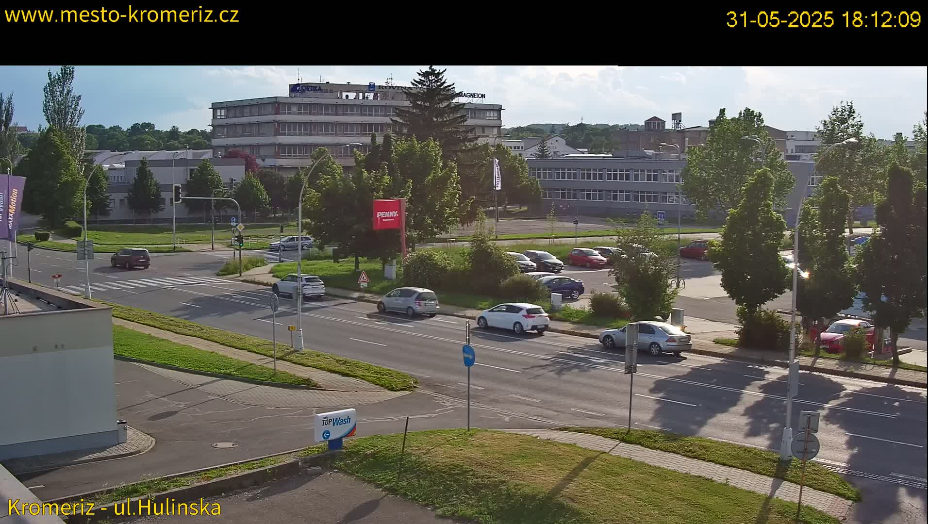 A sunny afternoon street scene shows several cars driving and parked near multistory buildings and lush green trees.