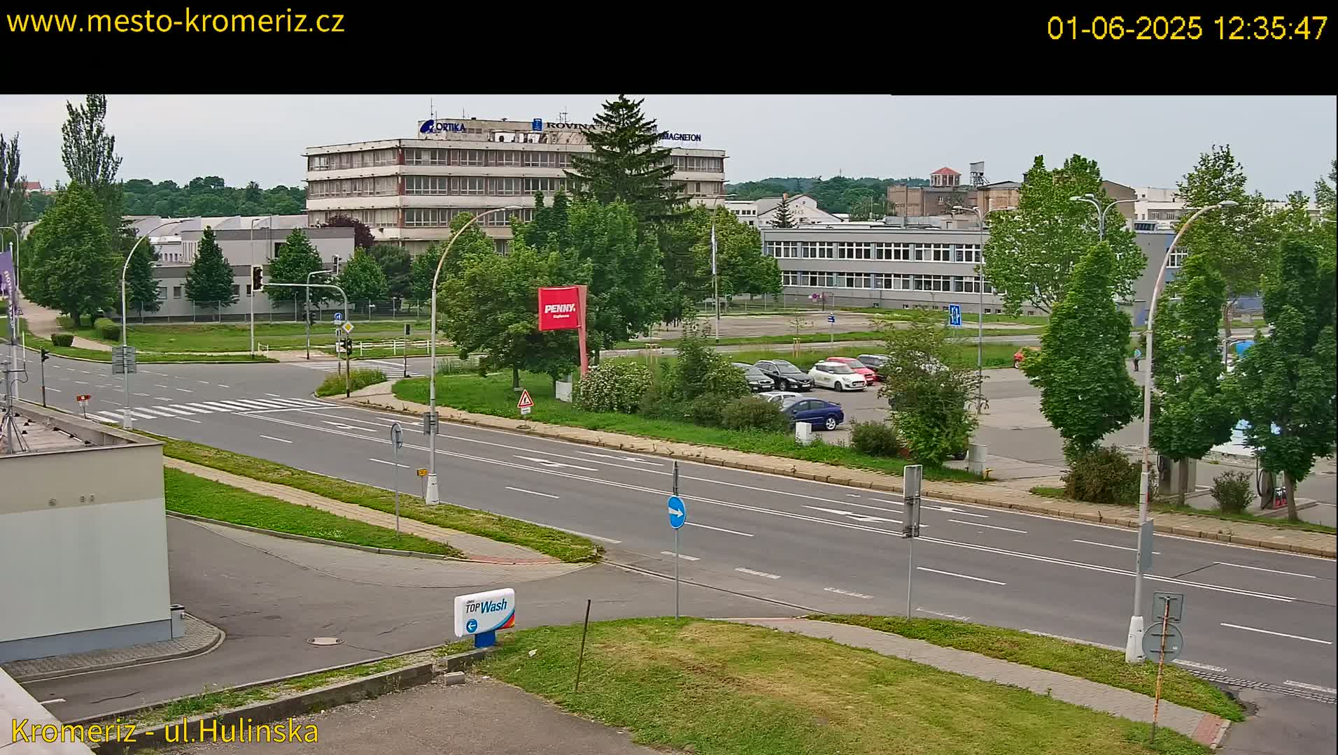 A mostly sunny day view of a multi-lane road, with several cars parked in a lot next to a large building, and other buildings in the background.