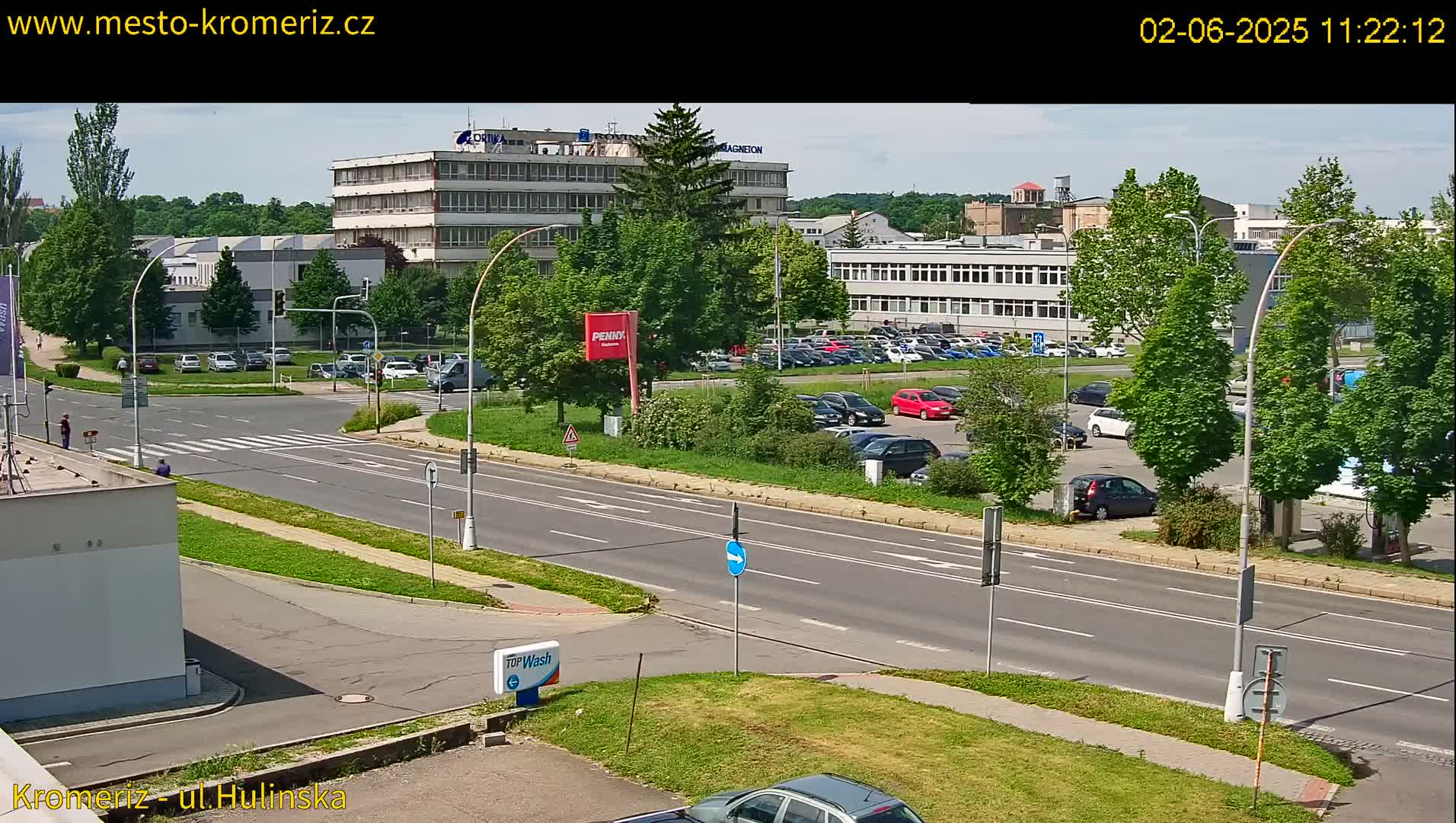 A sunny day reveals a multi-lane road with parked cars in a lot adjacent to several buildings, including a large industrial structure.