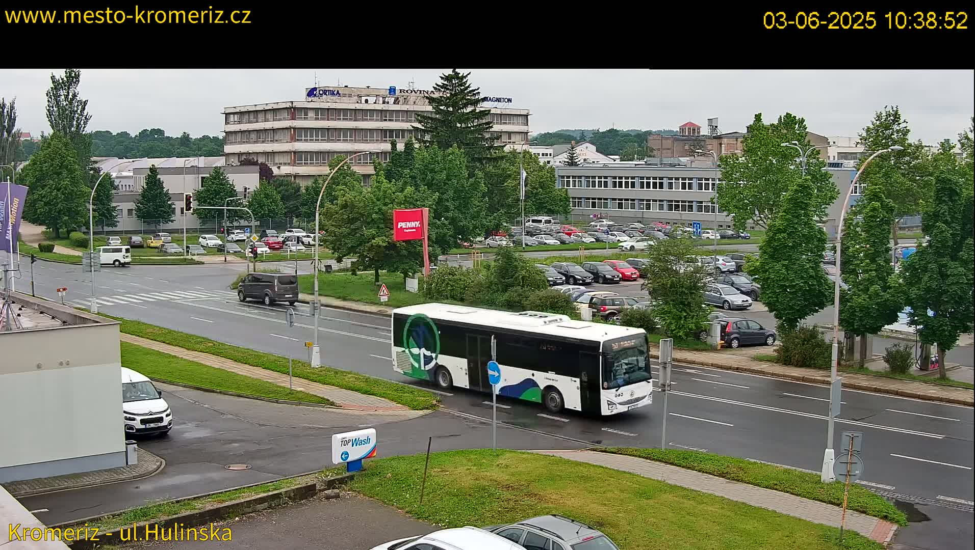 A white bus drives down a wet street past a parking lot and several buildings on an overcast day.