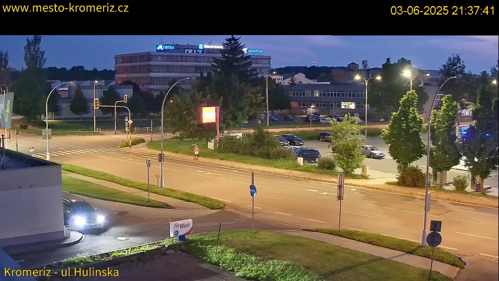 A nighttime, illuminated street scene shows a multi-lane road with cars, streetlights, and a parking lot with cars parked in it next to a building.