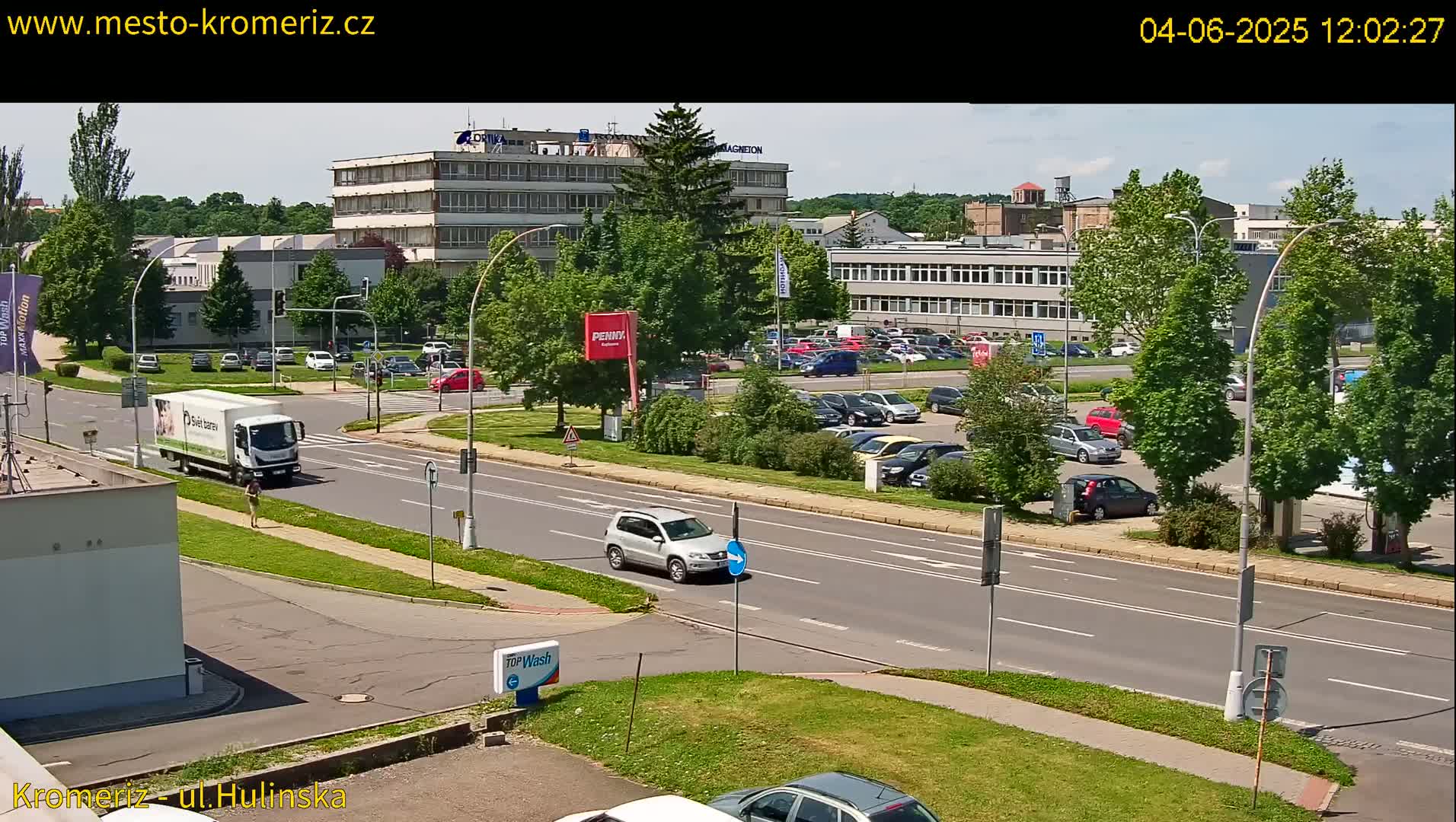 A sunny day reveals a city street scene with numerous parked cars in a lot, a few moving vehicles on the road, and several buildings visible in the background, all surrounded by lush green trees.