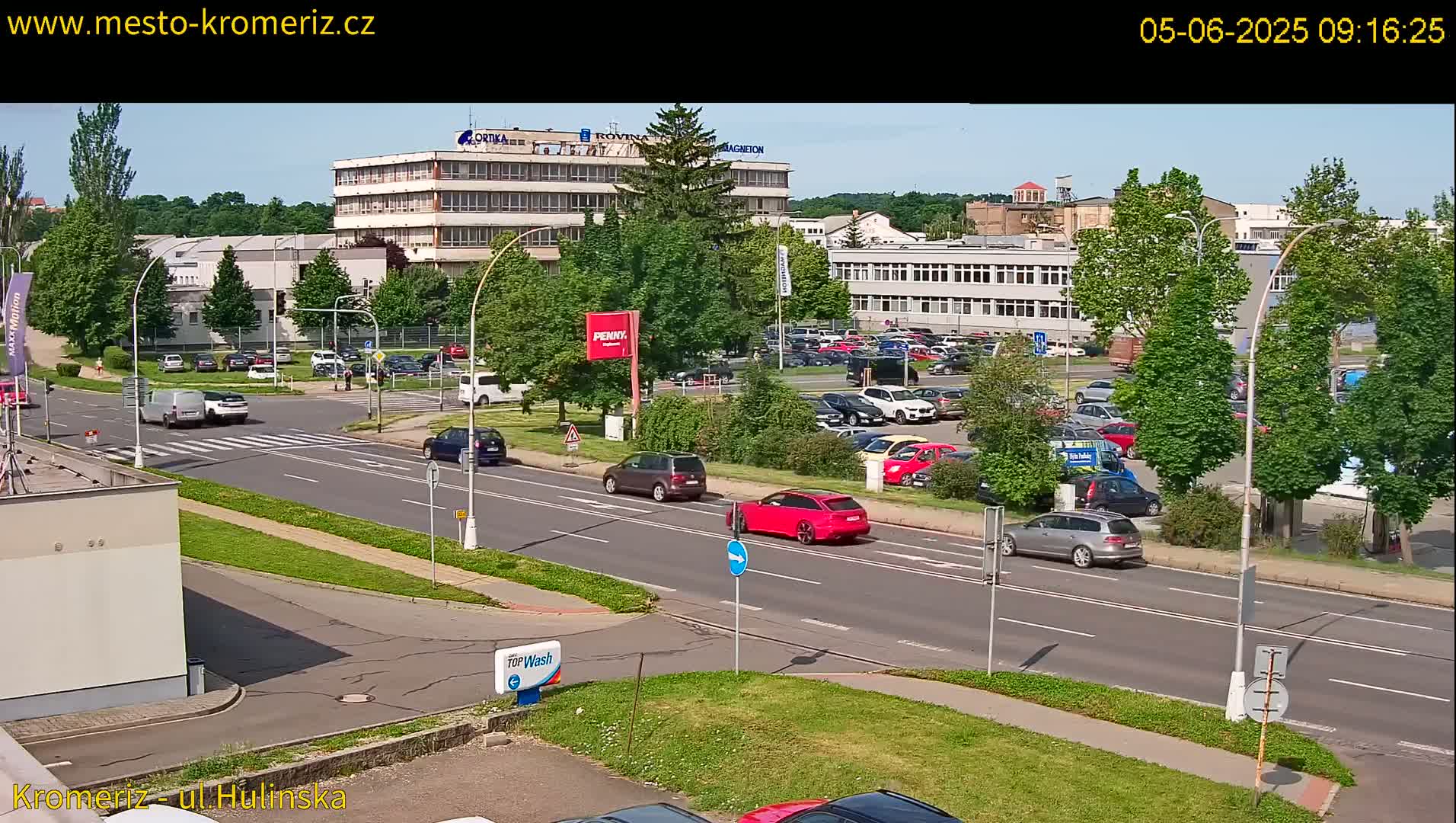 A sunny day reveals a multi-lane road with several cars driving past industrial buildings and a large parking lot filled with vehicles.