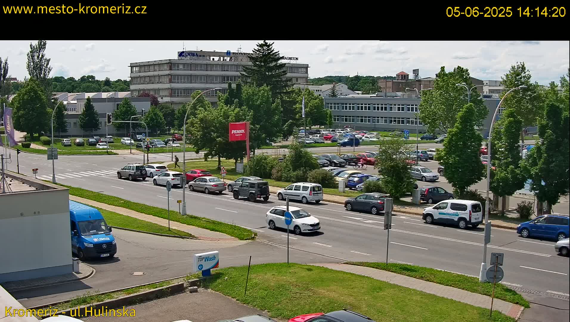 A sunny day reveals a multi-lane road with various cars driving past a large parking lot and several multi-story buildings.