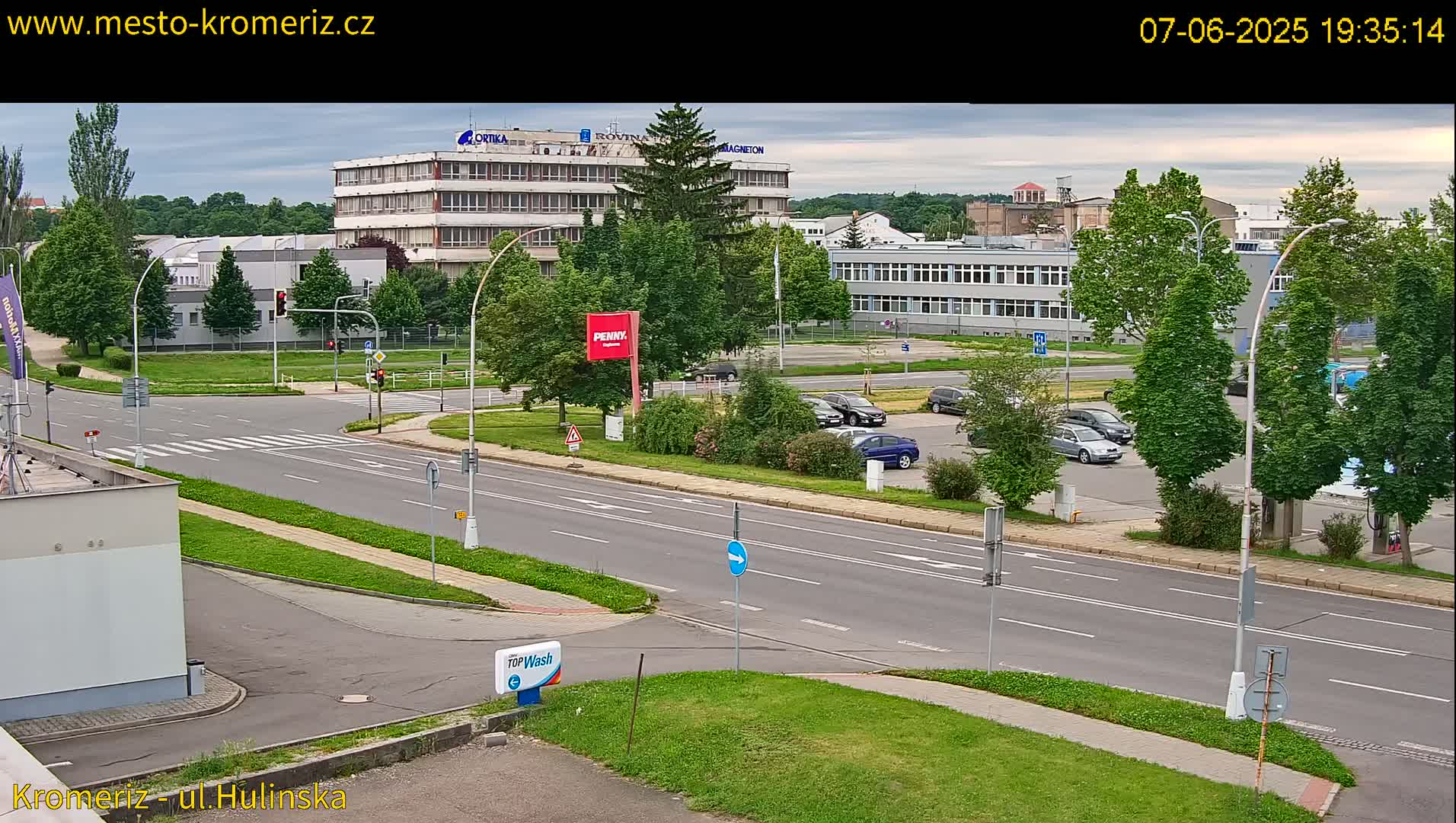 A mostly sunny, overcast street scene shows a multi-lane road with several cars parked in a lot next to a large building, surrounded by lush green trees and landscaping.
