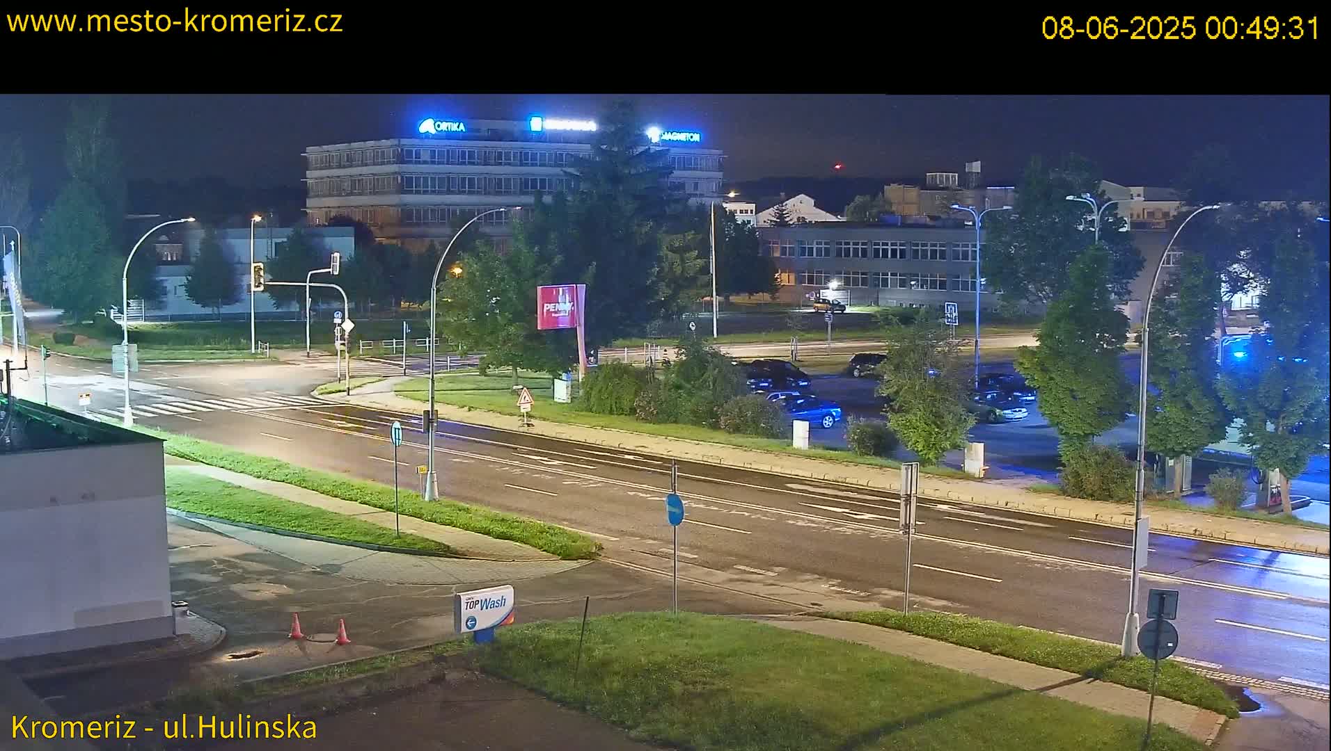 A nighttime, wet road scene shows a multi-lane road with cars parked in a lot and a few cars driving, near several buildings under bright streetlights.