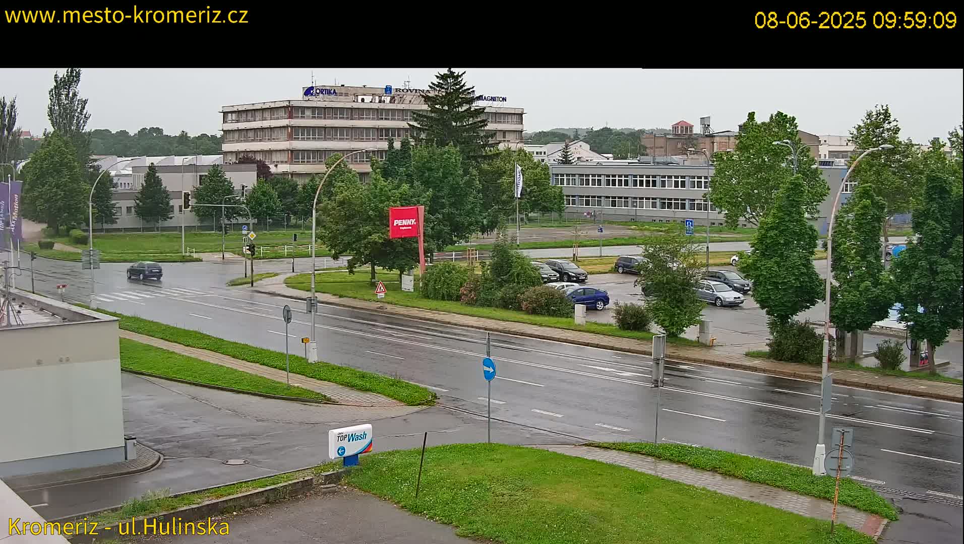 A wet, overcast day reveals a view of a multi-lane road with several cars parked in a lot and driving on the road, alongside industrial buildings and lush green landscaping.